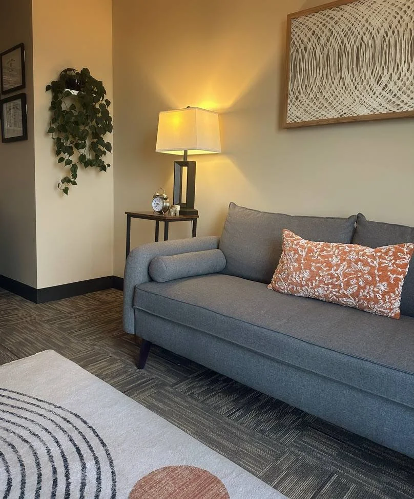 A cozy living room corner with a gray upholstered sofa, an orange floral cushion, a side table with a lit lamp, an alarm clock, and framed artwork on the wall.