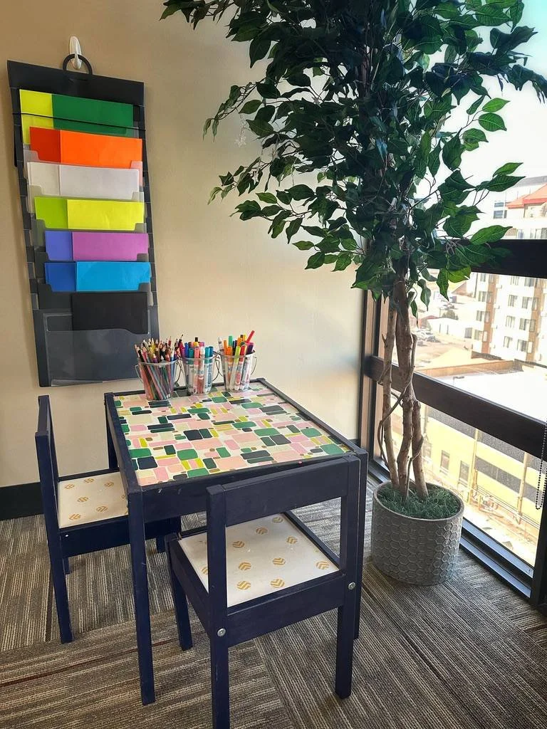 A small table with a geometric patterned tablecloth, surrounded by two chairs with patterned seat cushions, near a large window with a view of buildings outside. On the table, there are cups filled with colorful markers. Behind the table, there is a large potted plant and a wall-mounted organizer with colored folders.