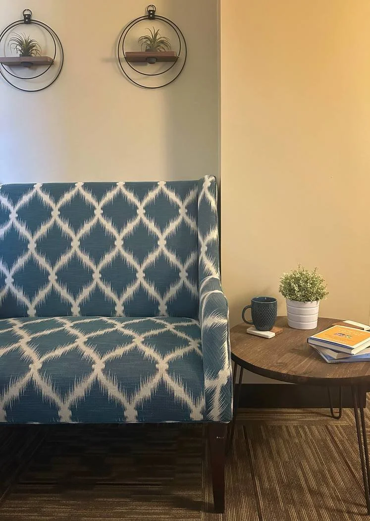 A cozy living space corner with a blue armchair featuring a white patterned fabric, a wooden side table holding a potted plant, a dark coffee mug, and a stack of books. The wall behind has two decorative round shelves with small plants.