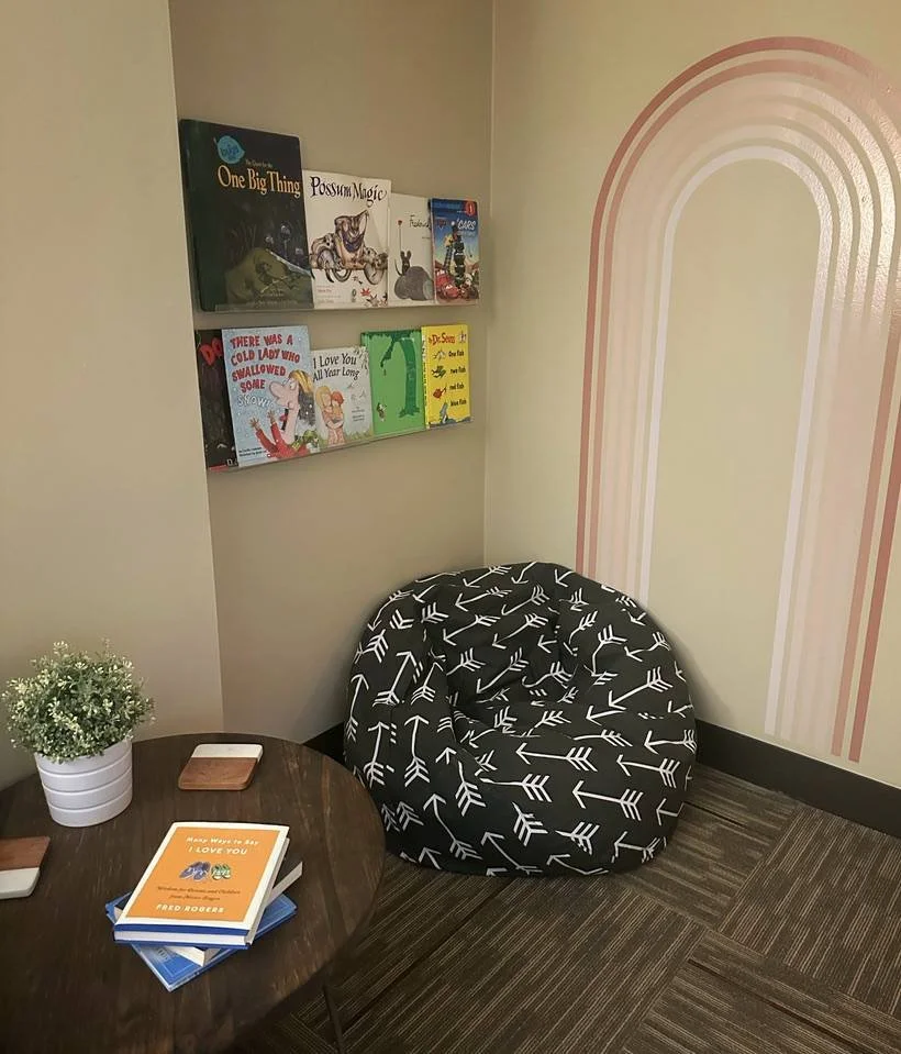 A cozy corner with a round wooden table holding a book titled "Many Ways to Say I Love You" by Fred Rogers, a potted plant, and two small coasters. Next to the table is a black bean bag chair with a white arrow pattern. Above the chair, a wall-mounted bookshelf displays children's books, including "One Big Thing," "Possum Magic," and other colorful titles. The wall behind features a pink and white rainbow mural.