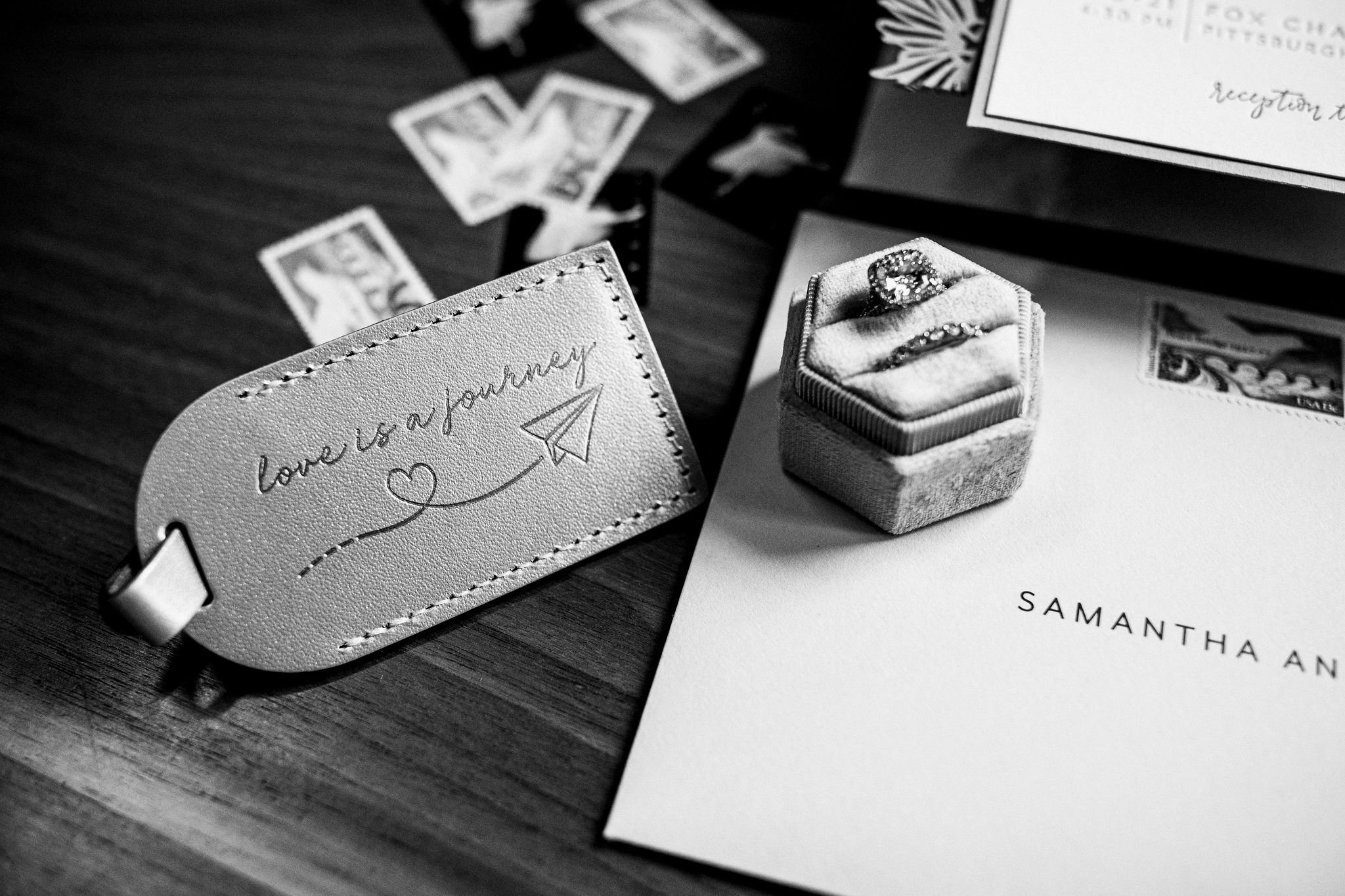 black and white photo of a leather luggage tag reading love is a journey next to a hexagonal velvet ring box holding an engagement ring and wedding band with wedding stationery and stamps scattered around them