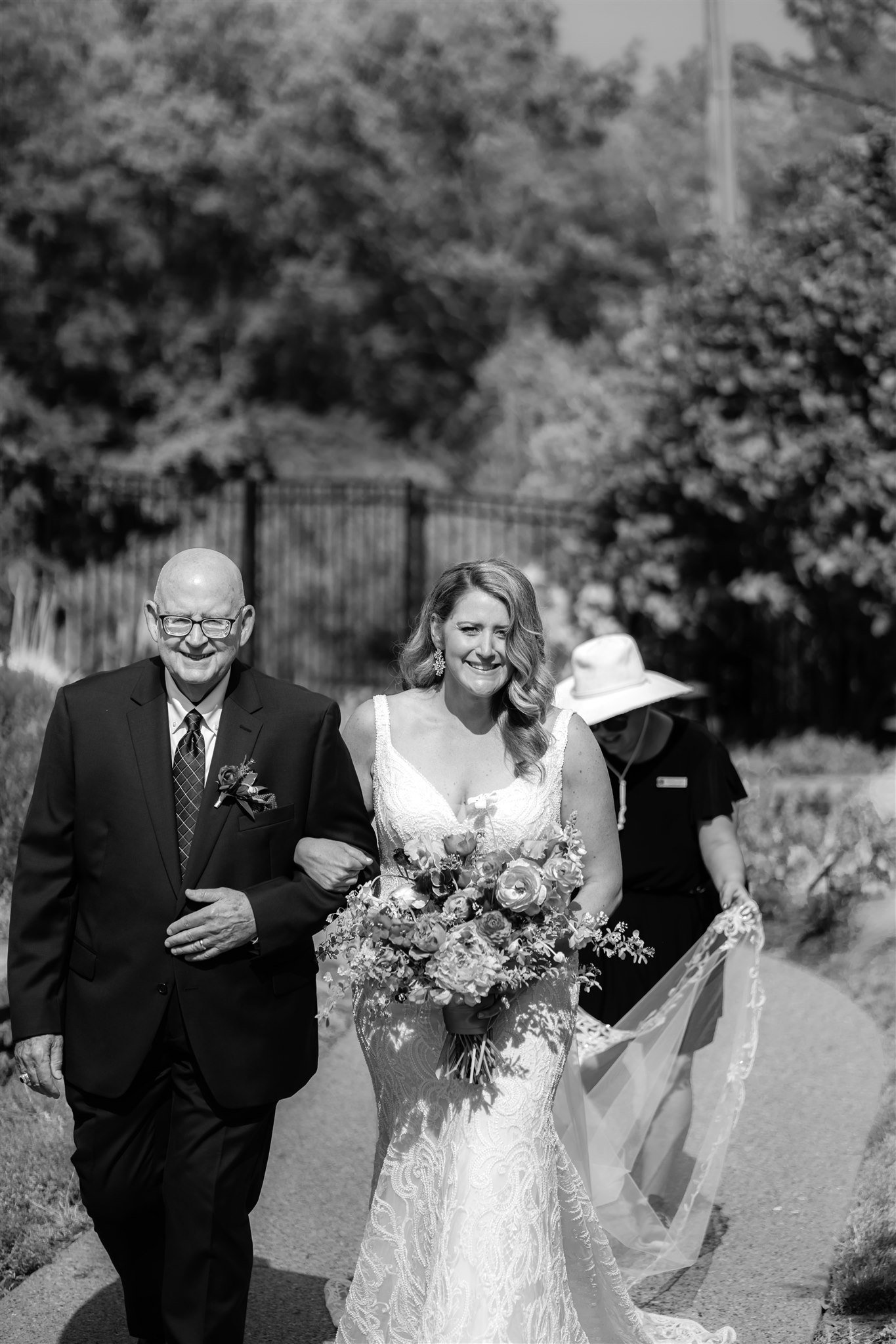 black and white photo of a bride and her father walking down the aisle at an outdoor wedding ceremony as a wedding coordinator carries the bride's veil and wedding dress train