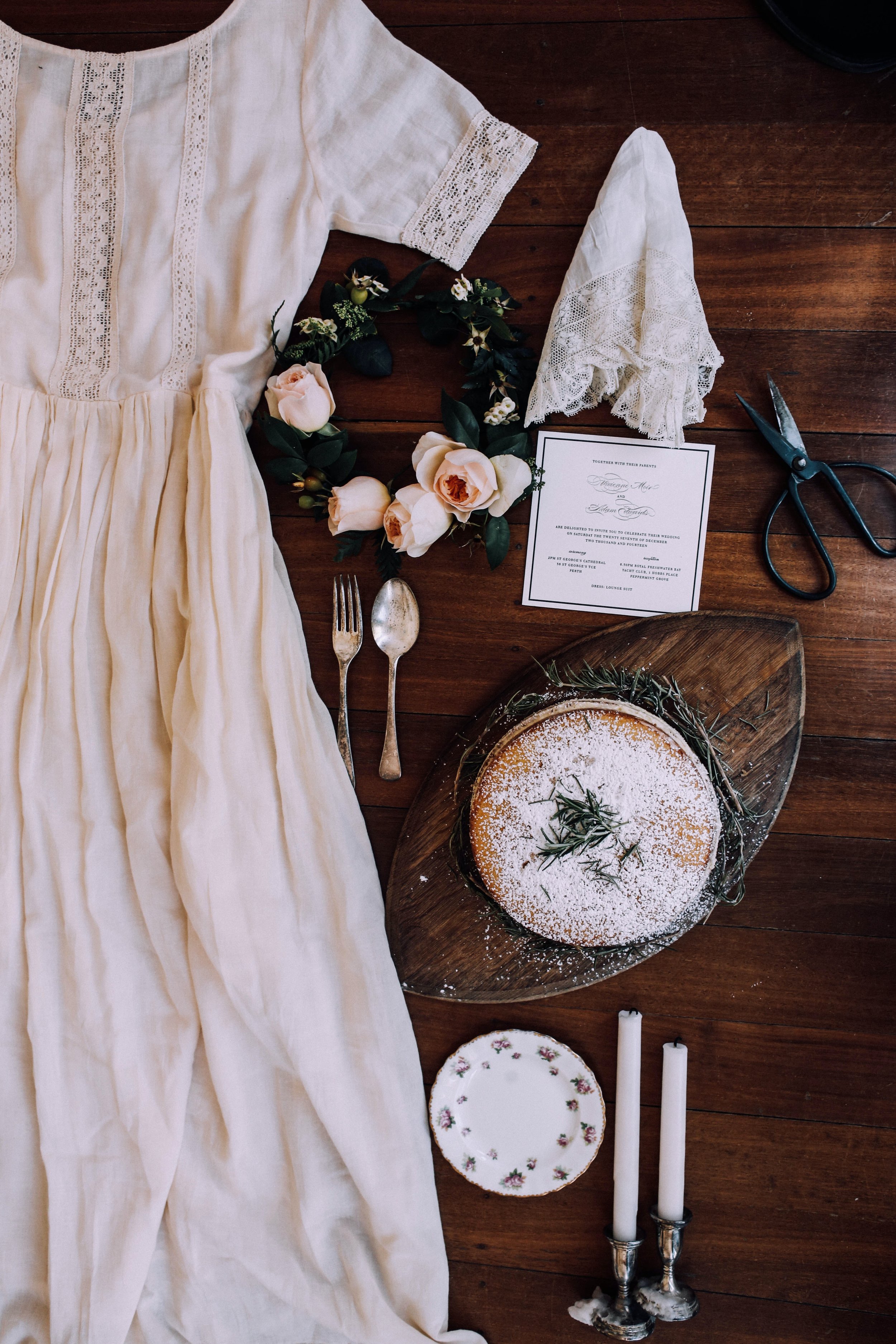 styleed flat lay on a wooden floor of vintage looking wedding dress and accessories with a rustic wedding cake and floral hair crown