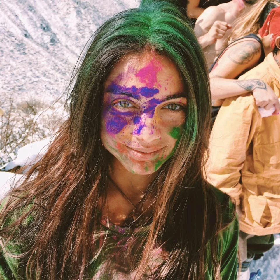 A woman with long brown hair, streaked with green, smiling with paint on her face in blues, pinks, and greens, surrounded by other people, some with tattoos, during a colorful outdoor event.