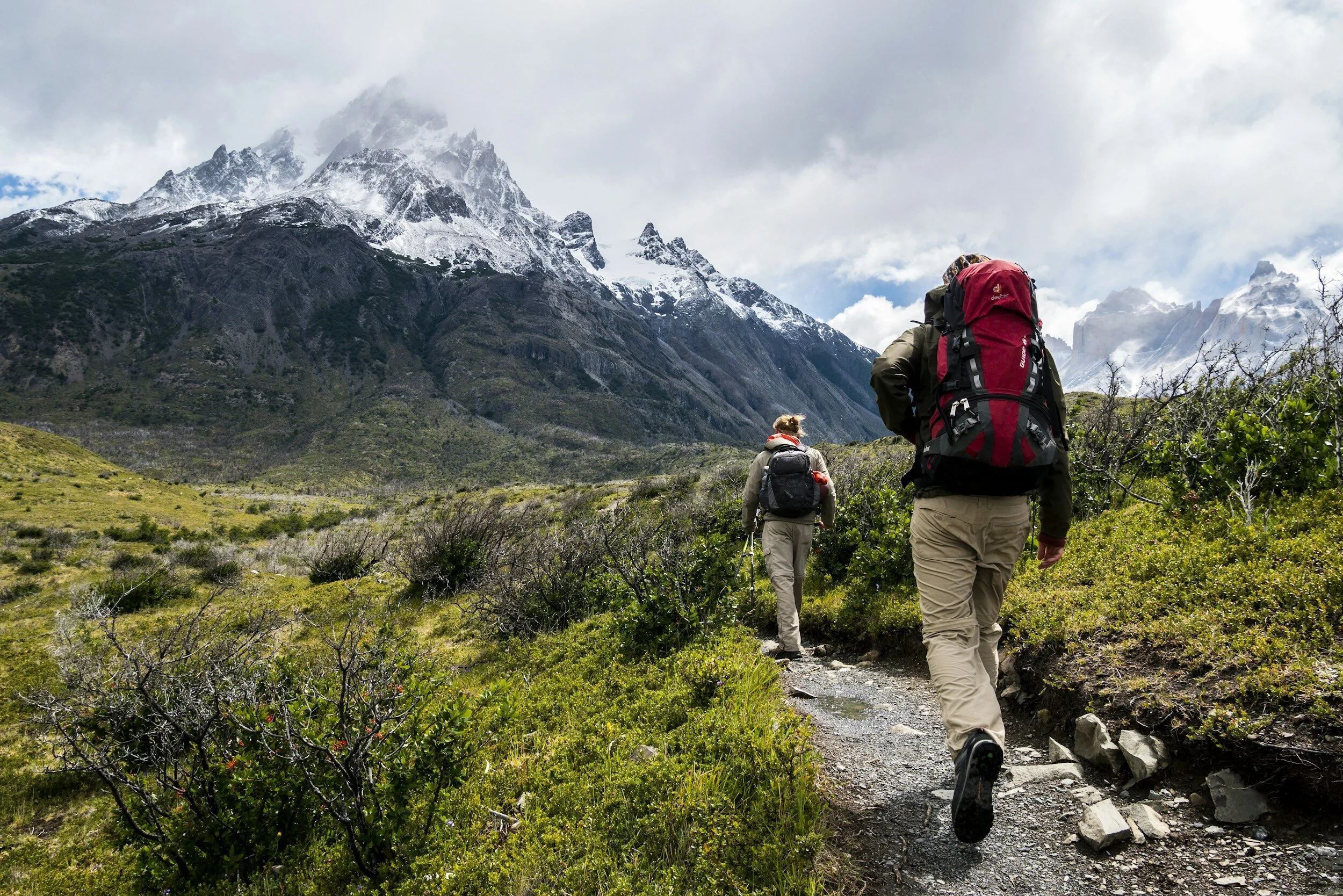 Two hikers with backpacks walking along a mountain trail in a lush green landscape with snow-capped peaks in the background.