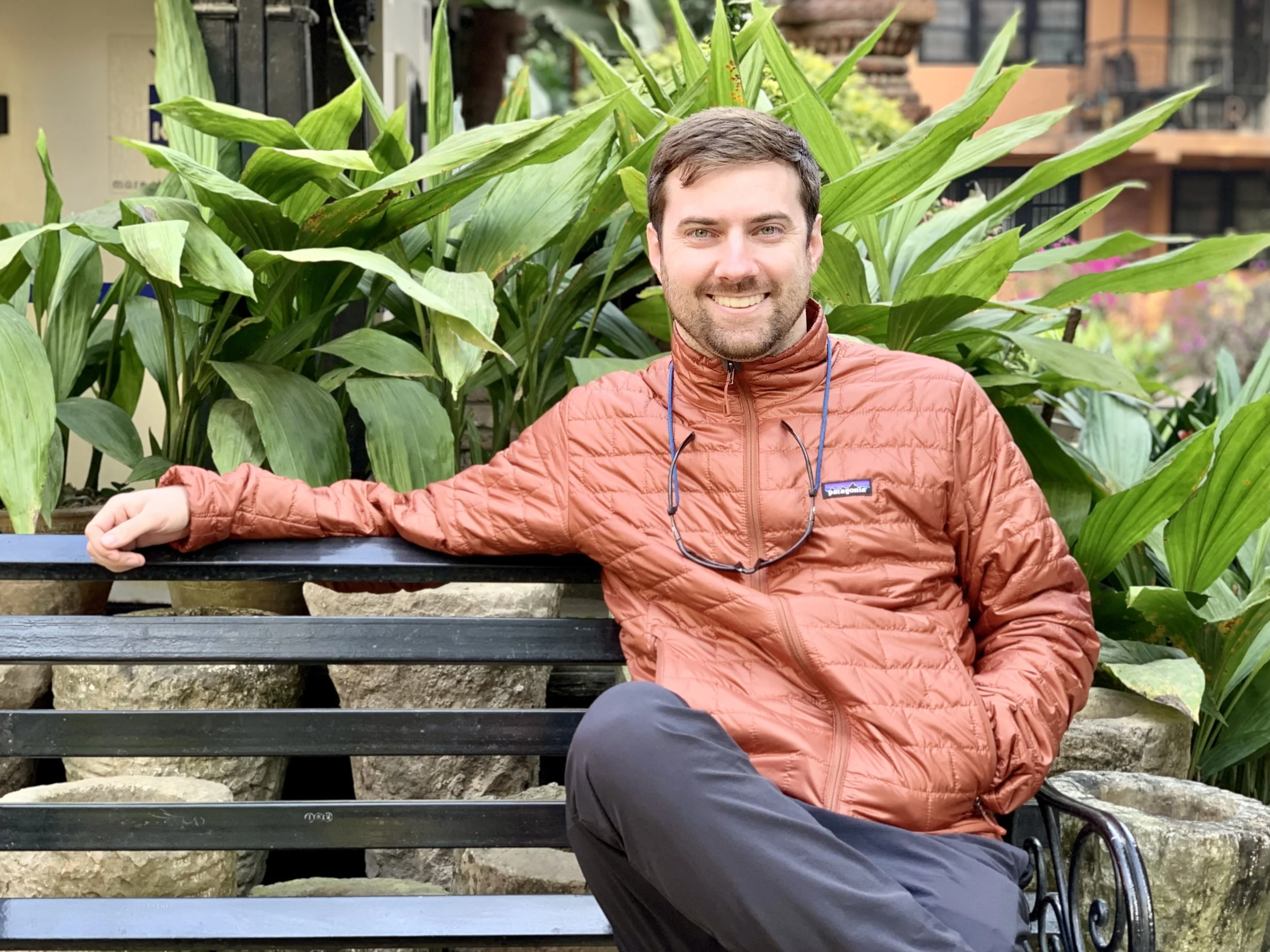 Man in an orange jacket sitting on a park bench with lush green plants in the background