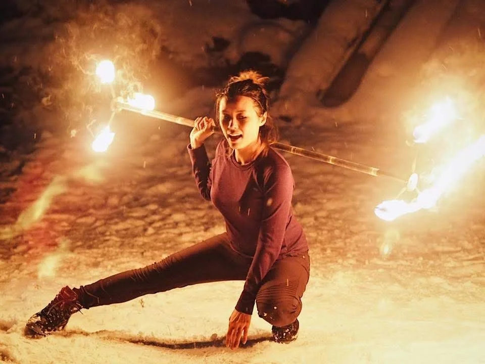 Woman performing a fire spinning routine outdoors at night, holding flaming poi or staffs, with snow on the ground.