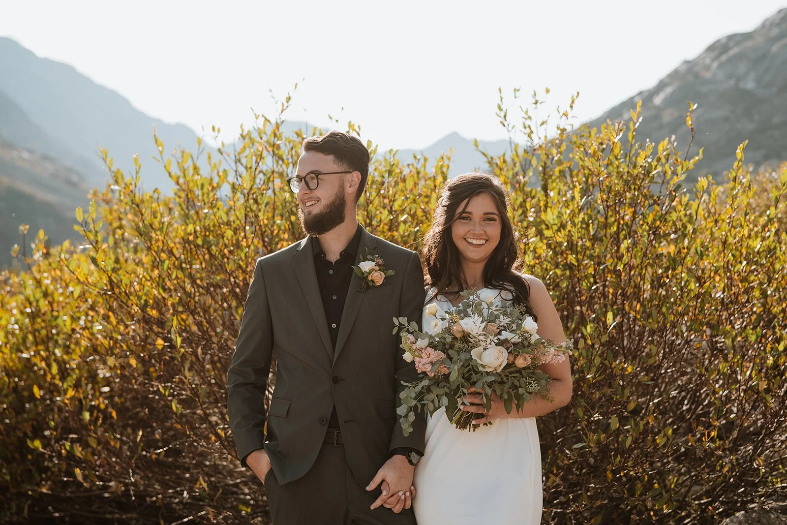 A smiling couple stands in front of a golden shrub with mountains behind them.