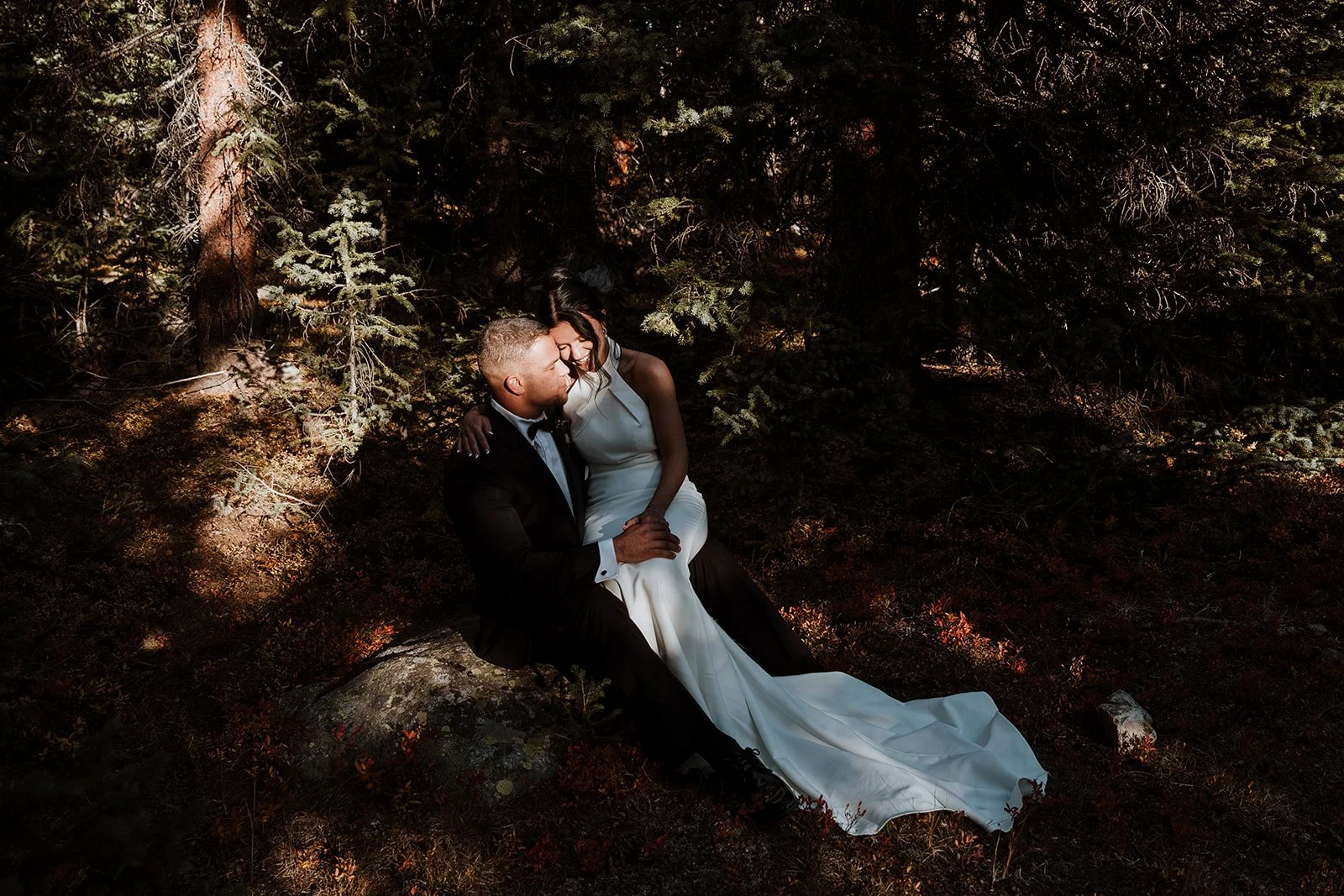 A bride & groom sitting together outside in nature while the sunlight shines on their faces.