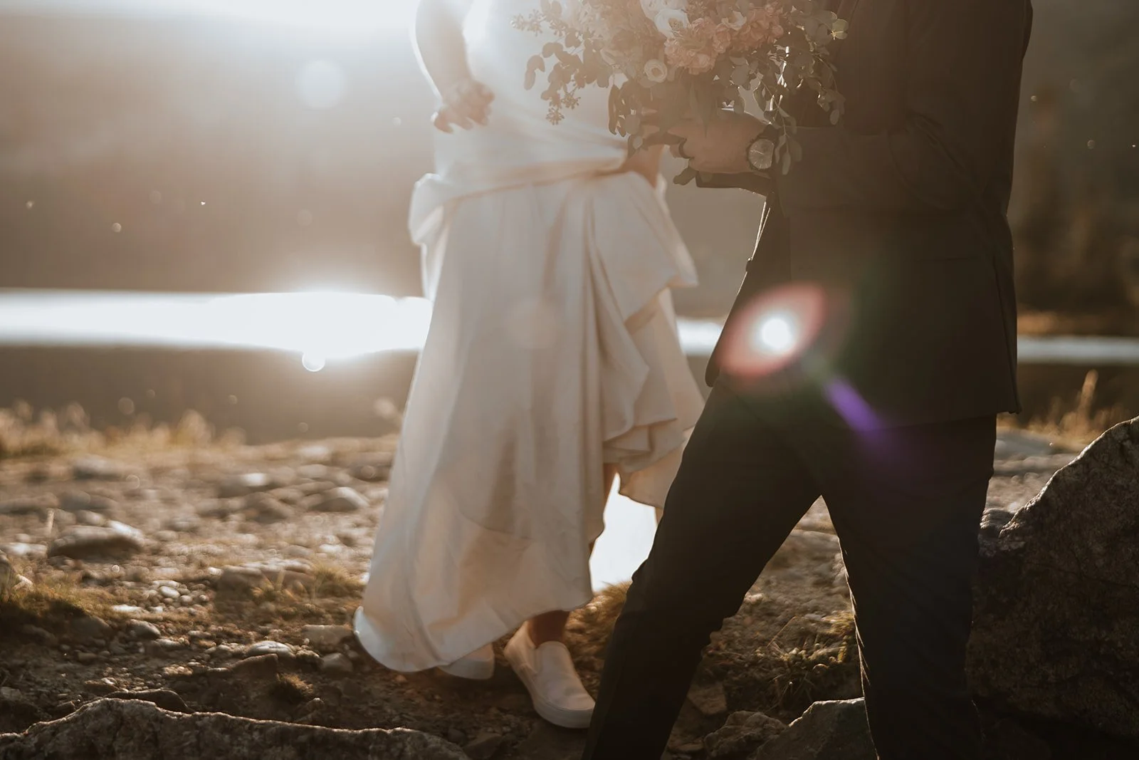 A bride & groom walking down a path with the sun shining on the lake behind them.