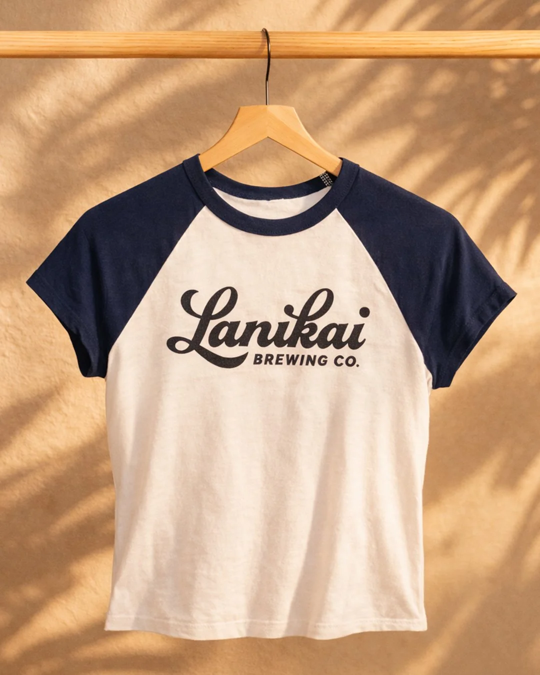 A cream-colored T-shirt with dark blue raglan sleeves and the text 'Lanikai Brewing Co.' printed in black on the front, hanging on a wooden hanger against a beige background.