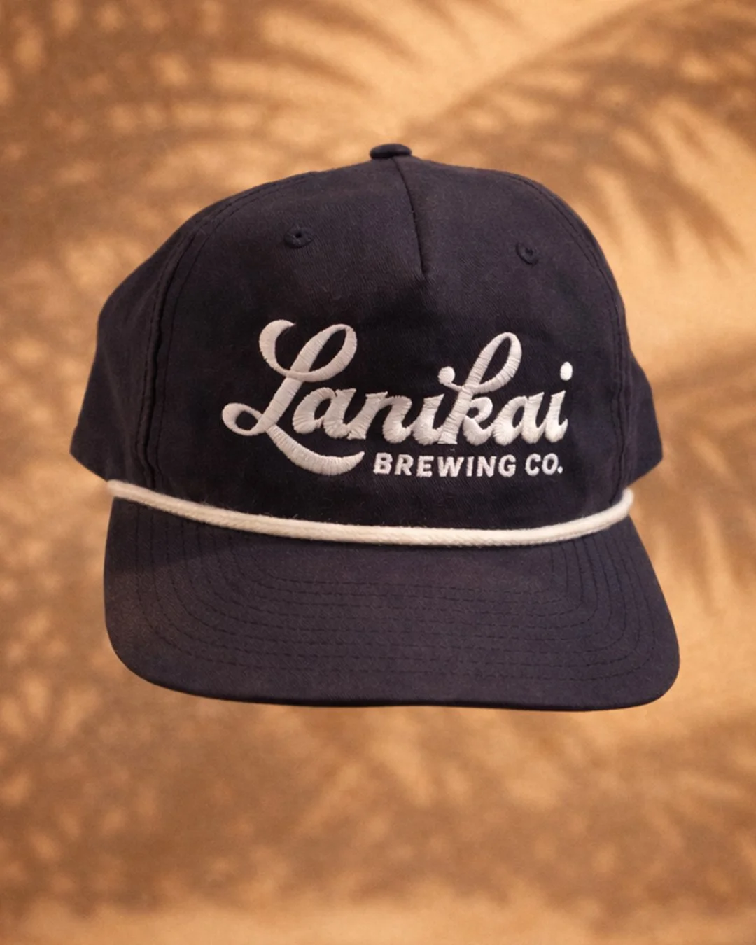 A black baseball cap with white embroidered text that reads 'Lanikai Brewing Co.', featuring a white edge around the brim, on a blurred brown background.