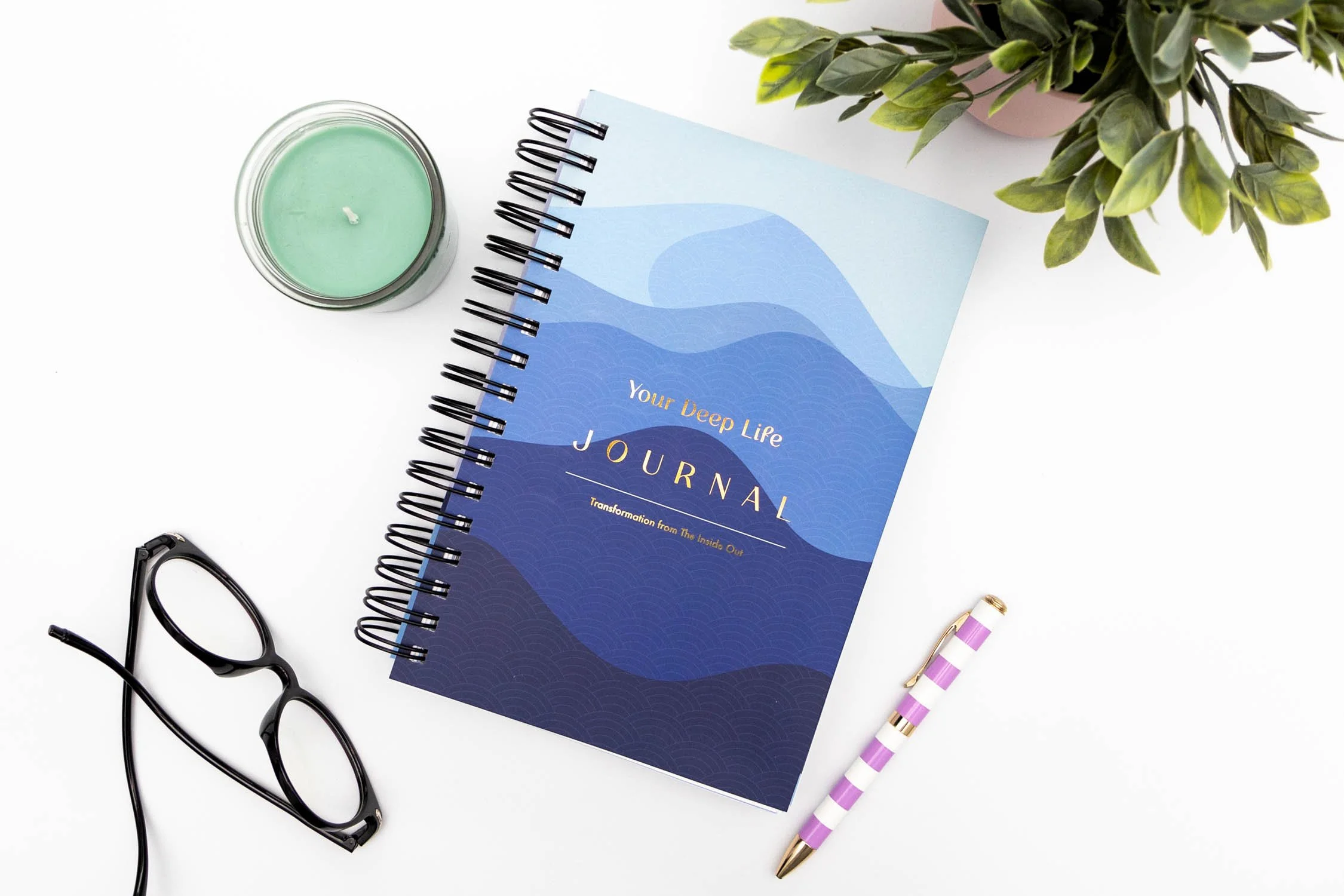 A journal titled "Your Deep Life" on a white surface, accompanied by a pair of black glasses, a striped purple and white pen, a green candle in a glass jar, and a green leafy plant in a pink pot.