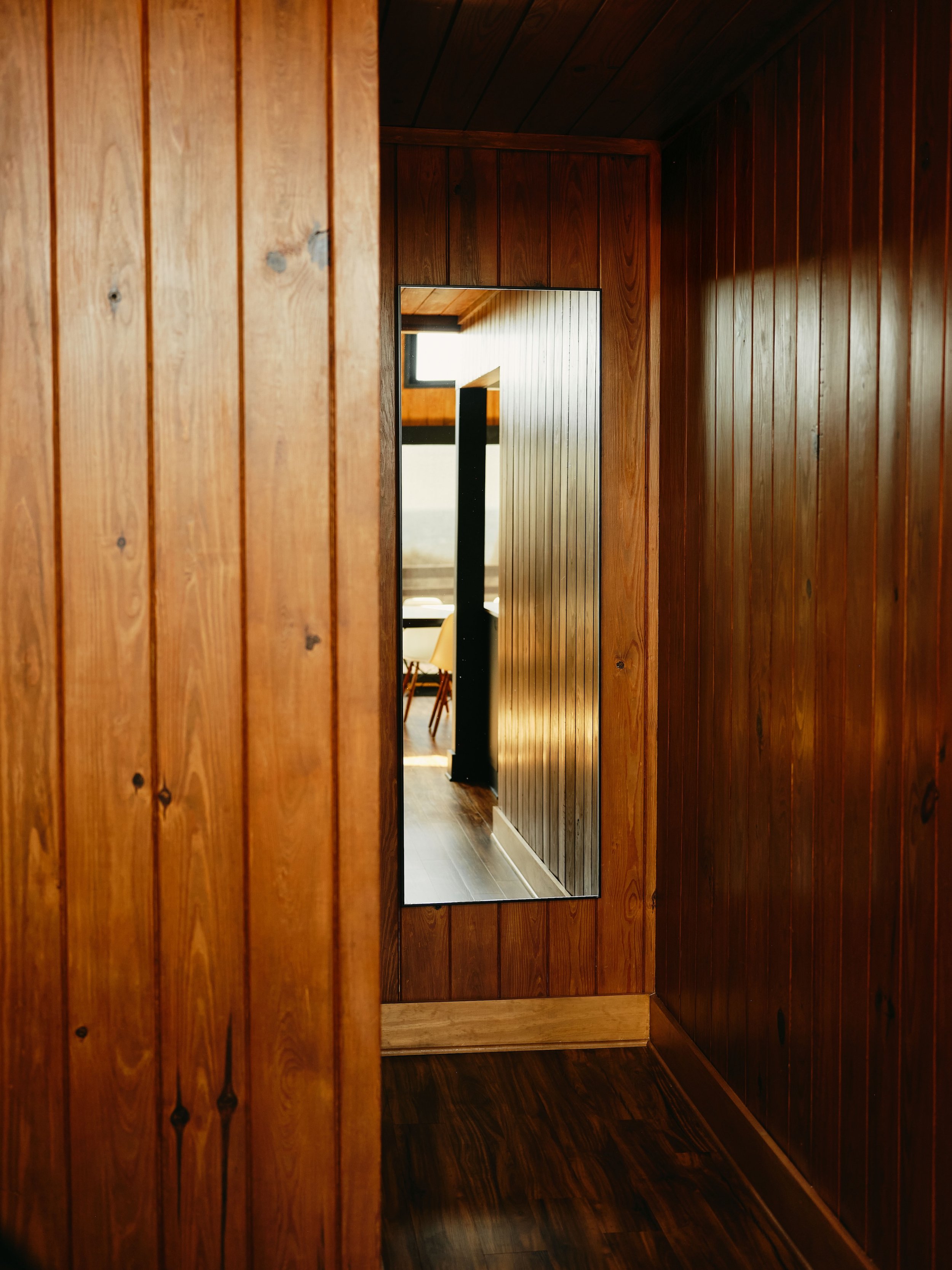 Wood-paneled hallway with a tall mirror reflecting a room with chairs and a window.