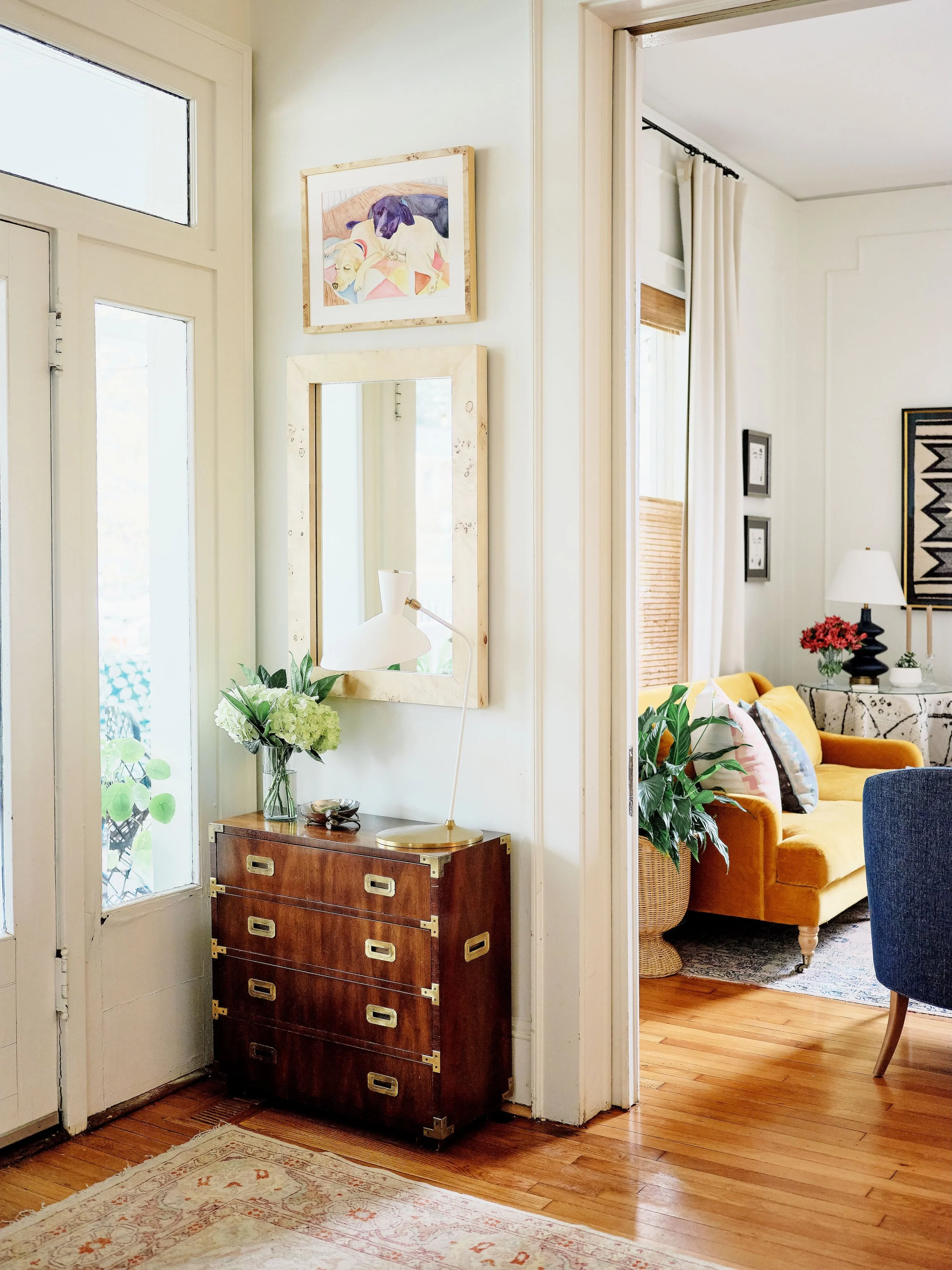 Interior view of a home entryway with a small wooden dresser topped with a plant, a vase with flowers, and a desk lamp. There is a mirror above the dresser and framed artwork on the wall. The adjacent living room contains a yellow sofa, a side table 