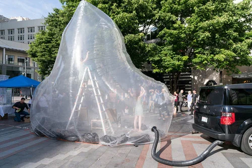 The Gas Trap - Coltura’s clear inflatable bubble connected to the tailpipe of a car. When the performance starts, the trap fills with smoke. Coltura is a nonprofit helping more people escape from the “gas trap”.