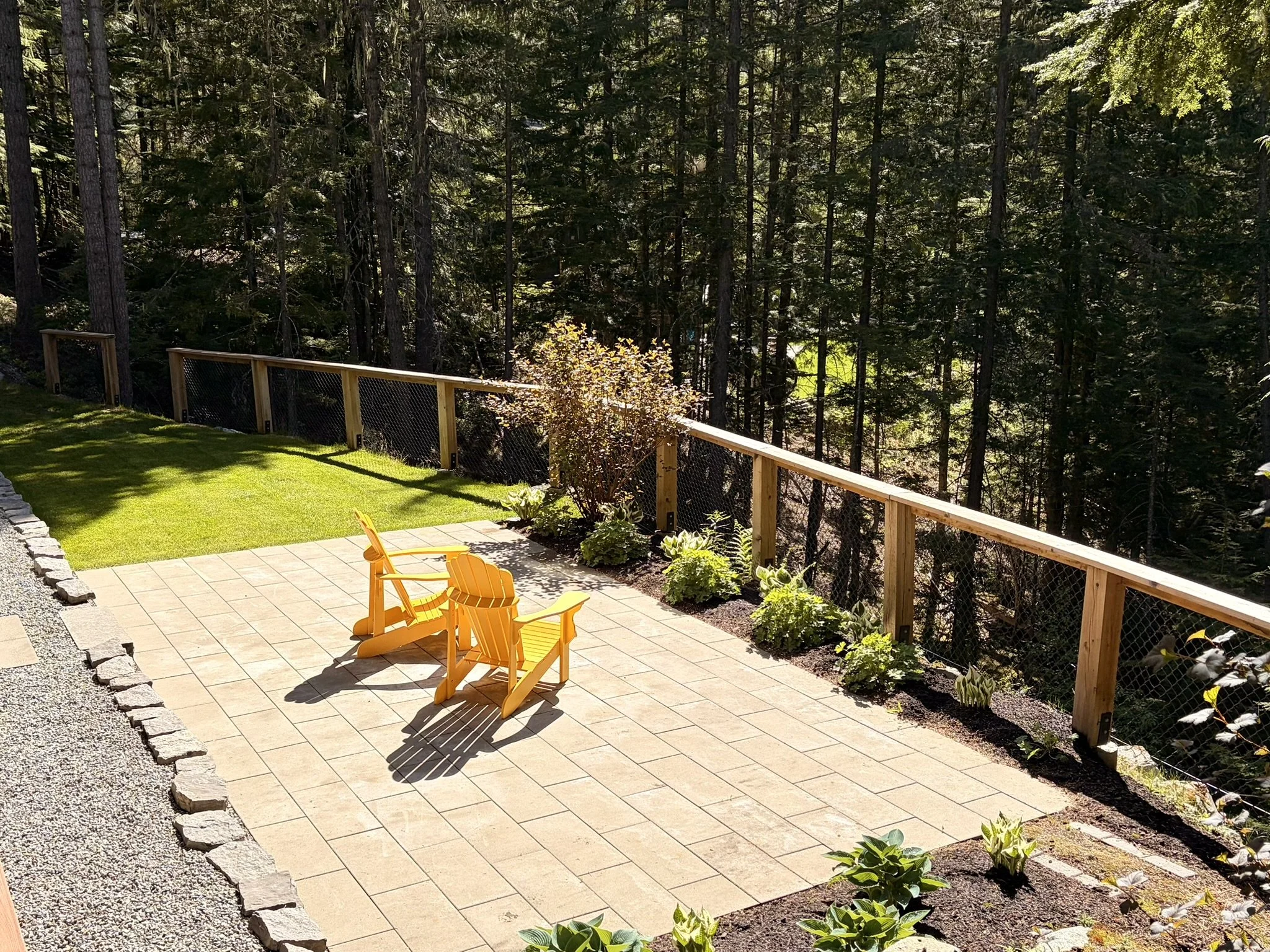 Patio area with two yellow chairs, bordered by garden beds with green plants, and a view of a dense forest in the background.