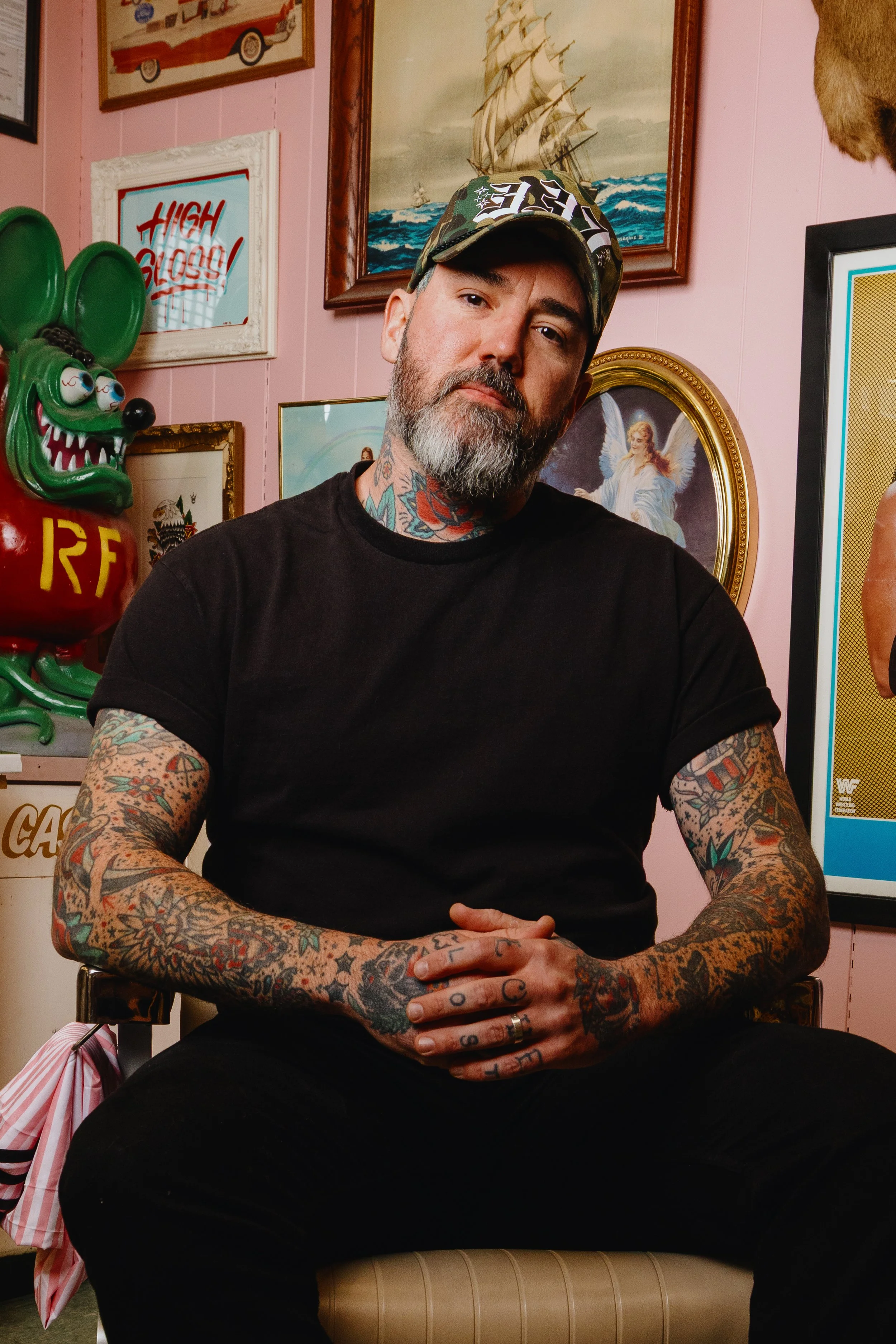 A man with a beard and tattoos wearing a black T-shirt and a camo cap sitting in front of a pink wall decorated with artwork and a green cartoonish mouse sculpture.