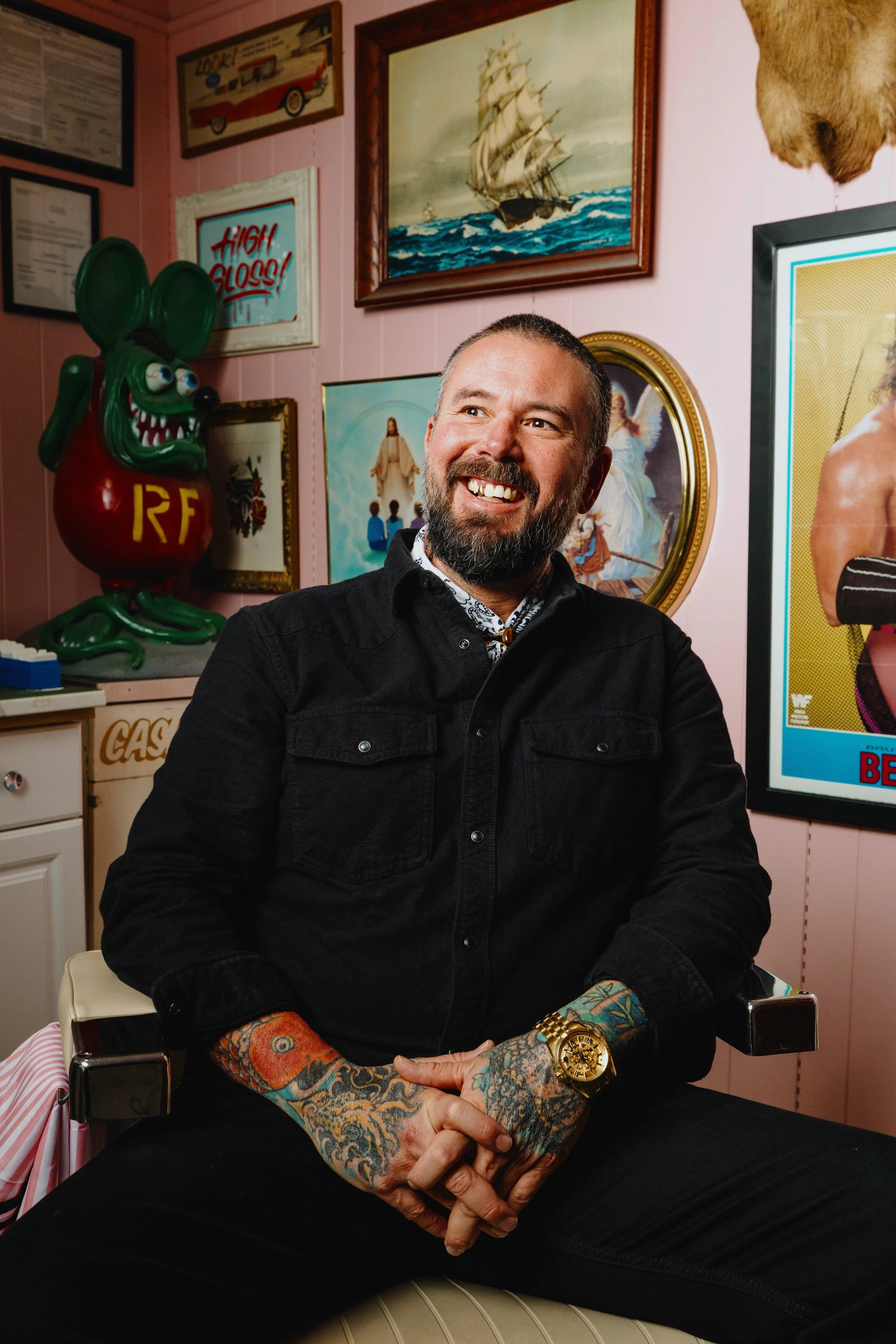 Man with tattoos and a gold watch sitting in a room decorated with artwork and signs, smiling and looking to his right.