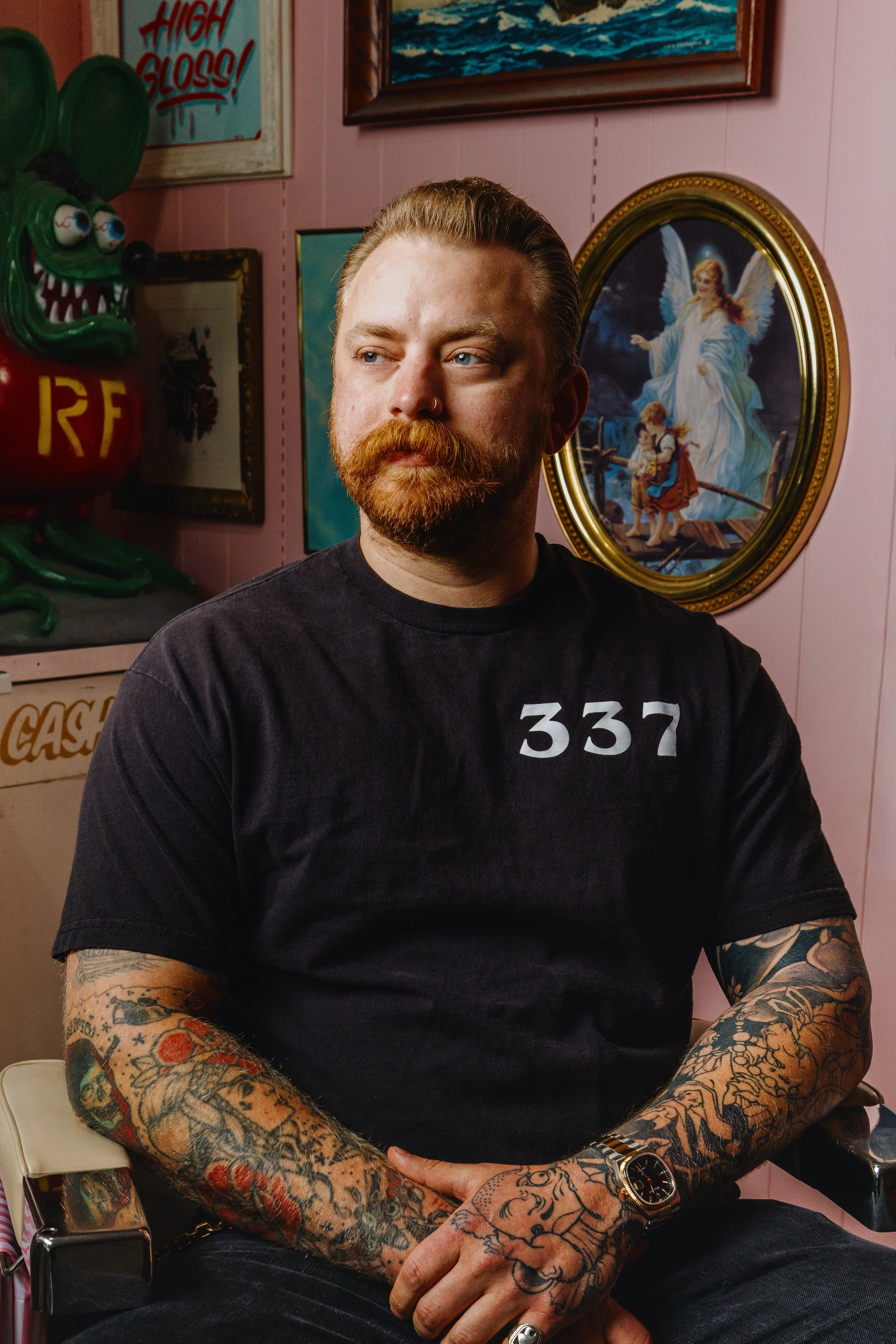 A man with red hair and beard, tattoos on arms wearing a black T-shirt with the number 337, sitting with hands crossed in front of him, surrounded by colorful framed pictures on pink wall.