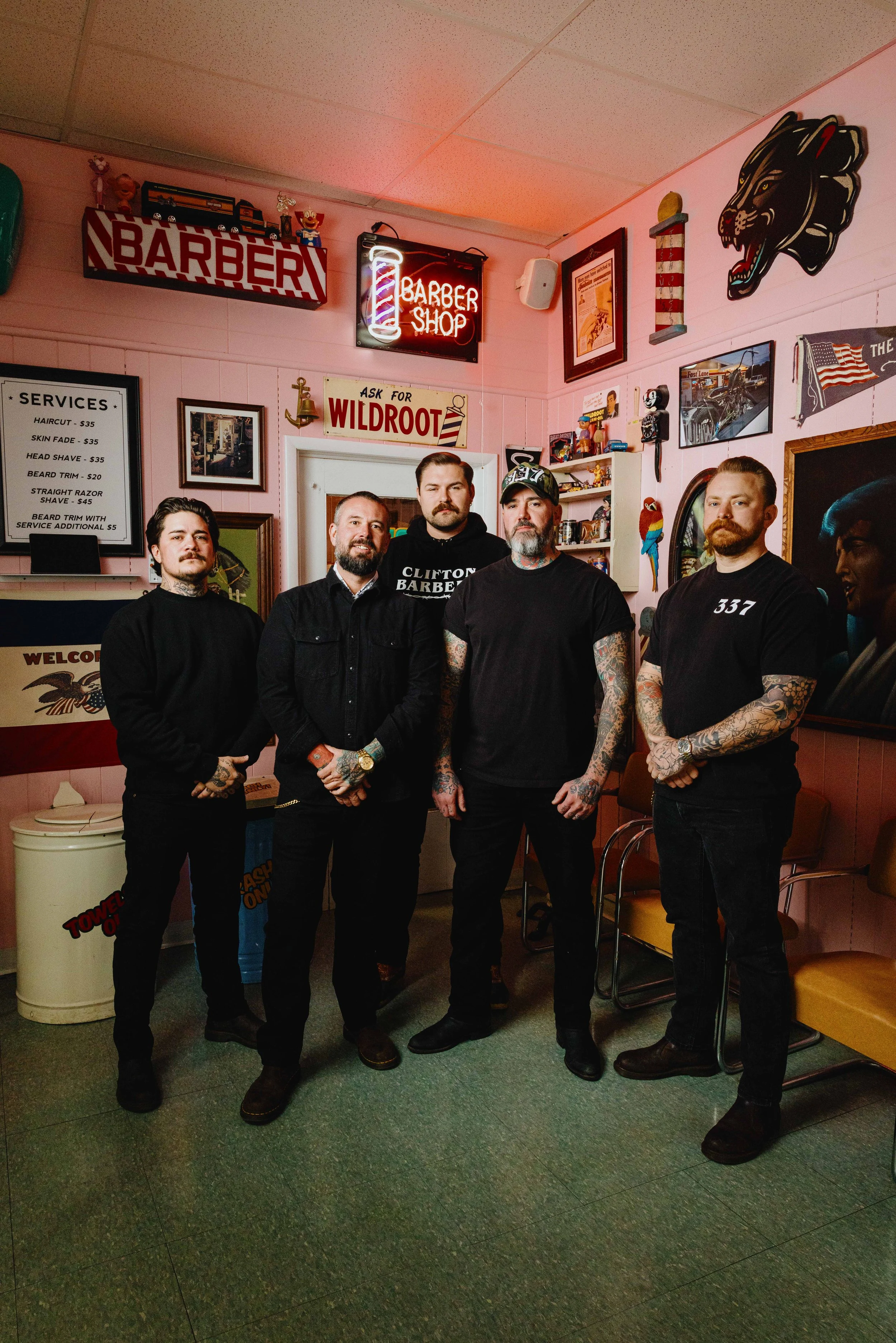 Five men standing inside a vintage barbershop with various signs and decorations on the walls.