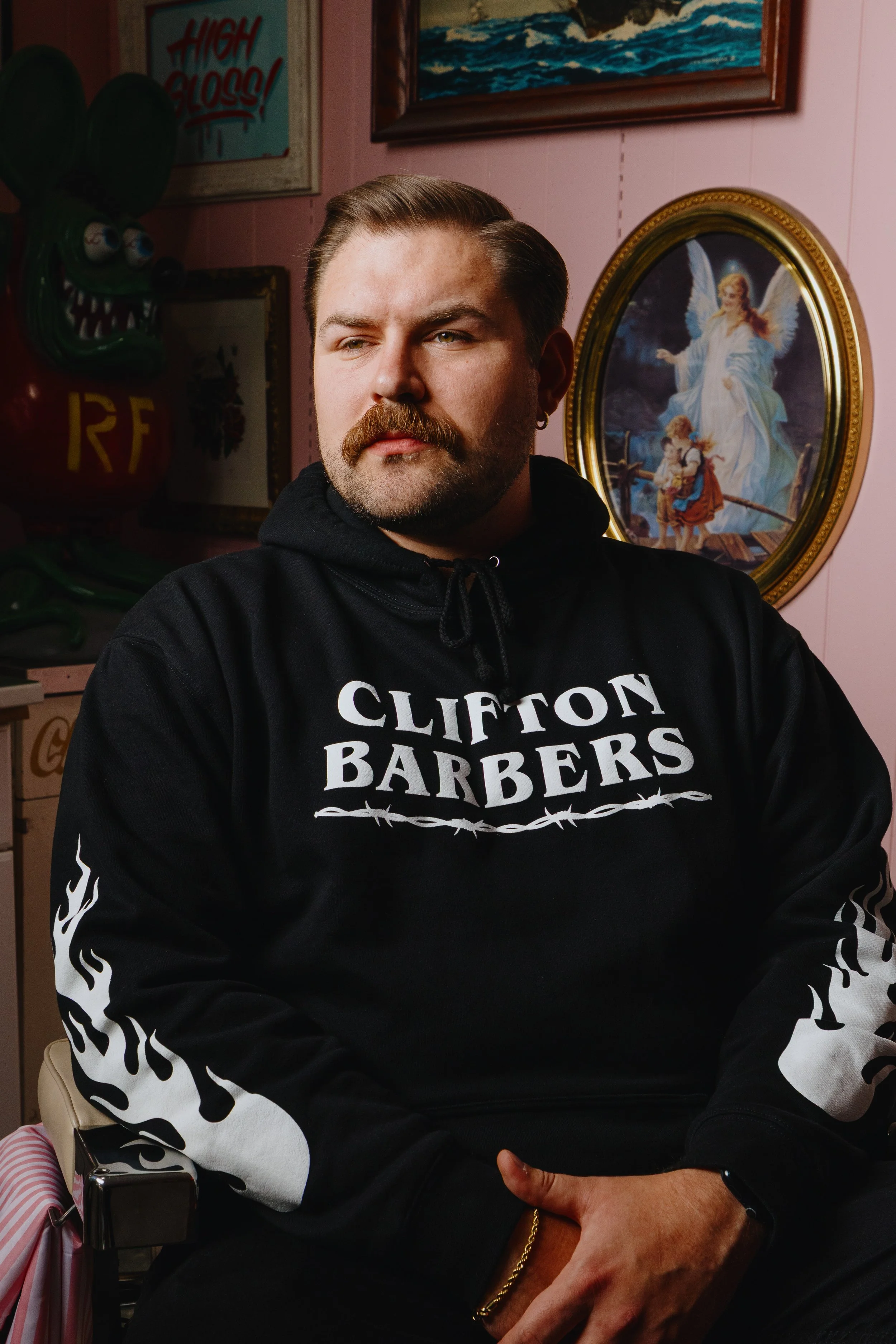 A man with a mustache and beard wearing a black hoodie with 'Clifton Barbers' logo sitting indoors with a riot of wall art in the background, including a painting of an angelic figure with children and a bright red and green balloon sculpture.