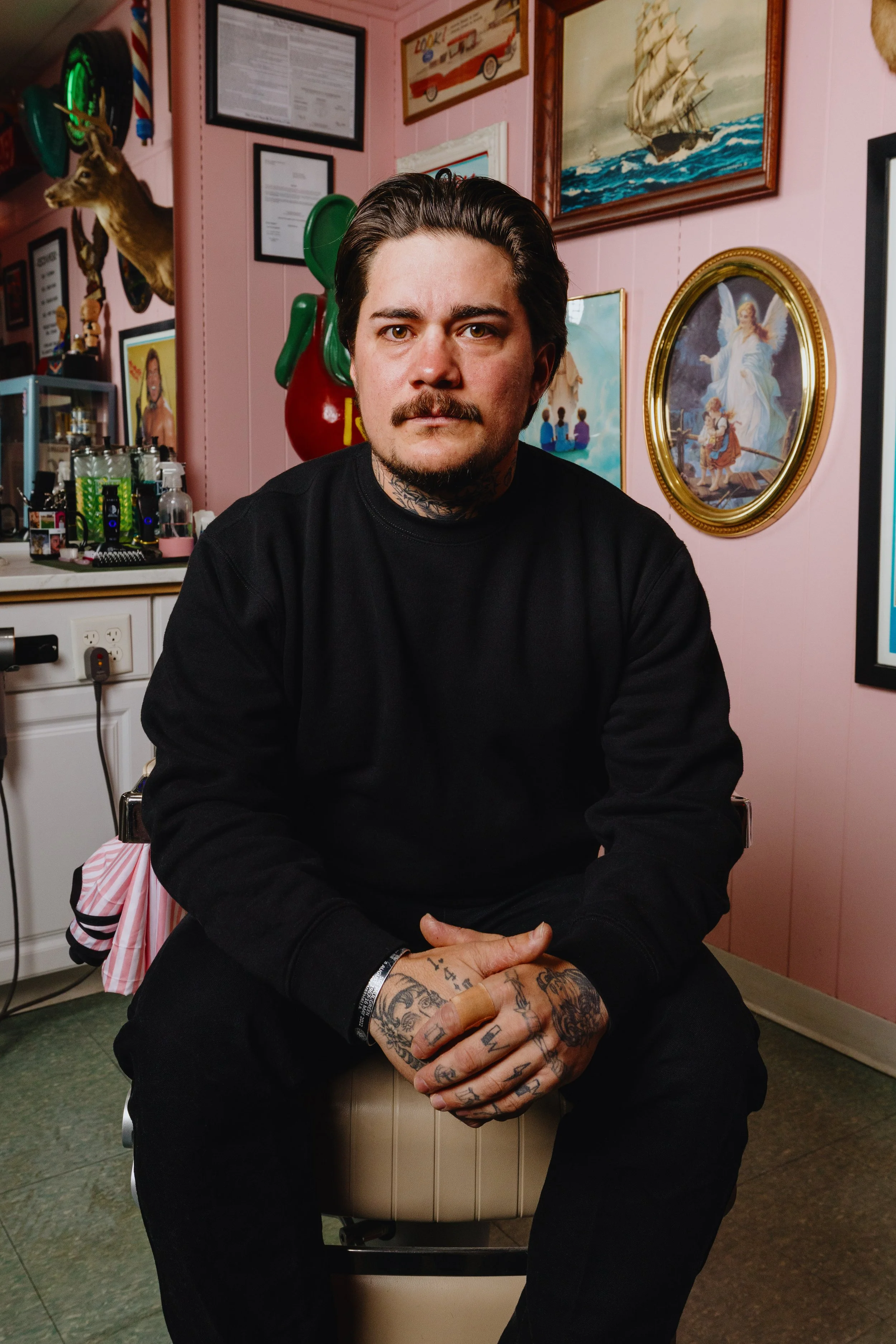 A man with tattoos and a beard, wearing a black sweatshirt, sitting on a stool in a room with pink walls decorated with various pictures and art, including a framed ship painting and religious artwork, surrounded by bottles and personal items.
