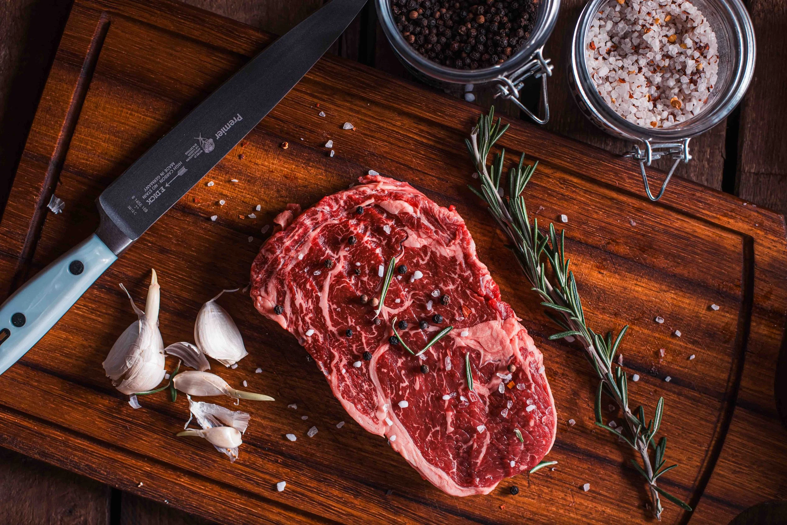 Premium ribeye steak på træskærebræt med groft salt, peberkorn, timian og skarp kniv ved siden af