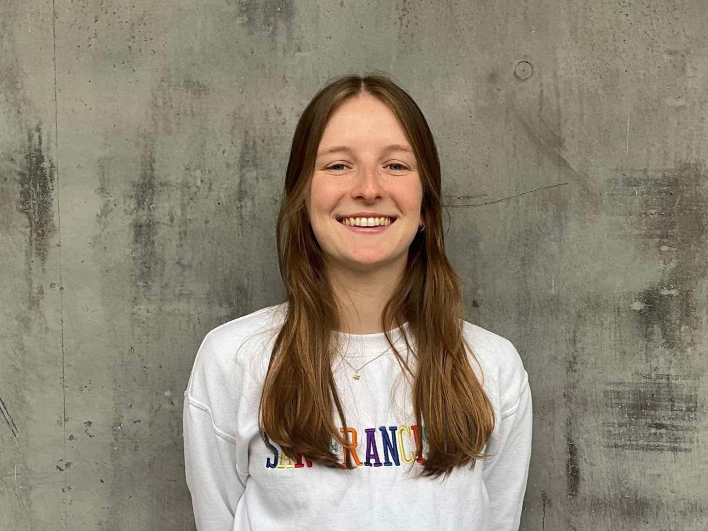 Psychologe Sigrid heet lang bruin haar, draagt een grijze sweater en poseert lachend voor een grijze muur.