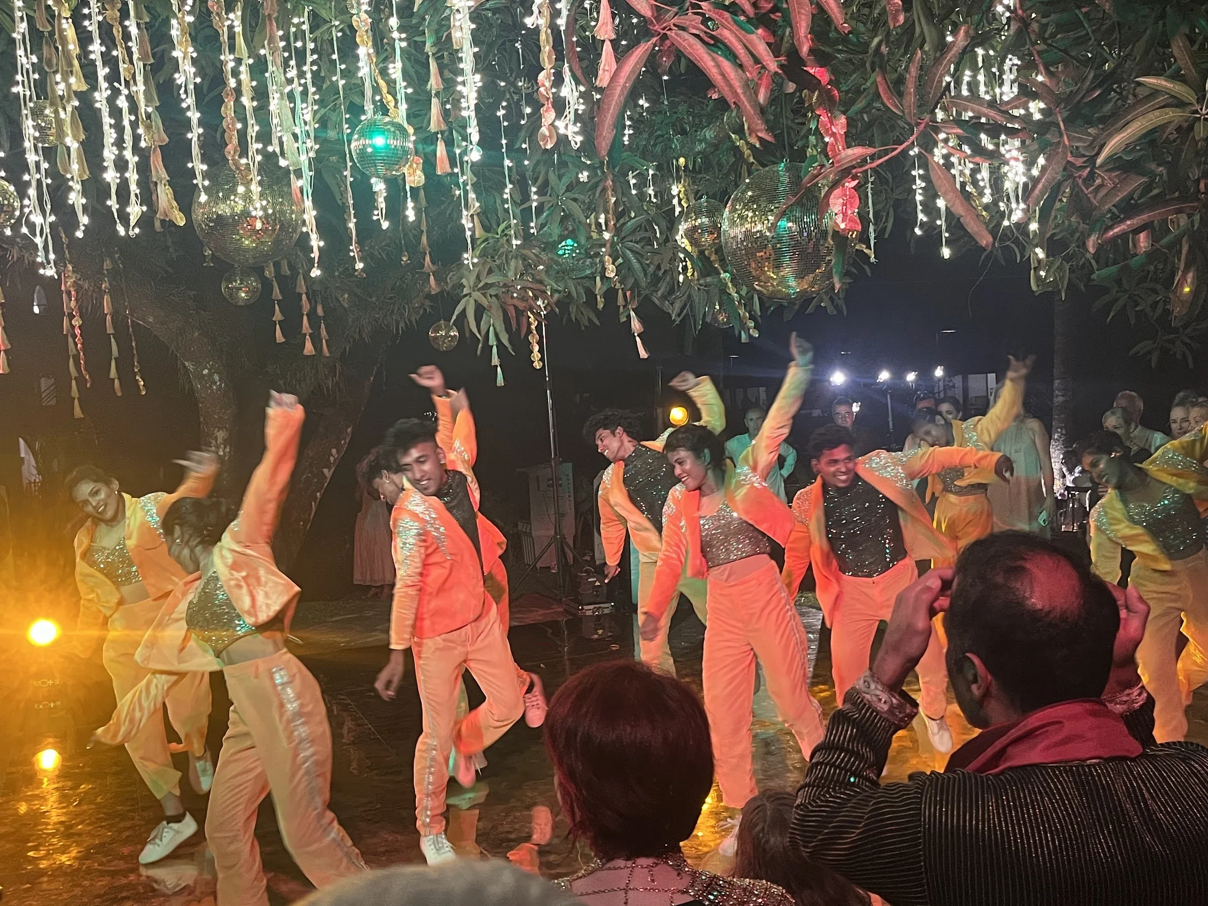 Bollywood dance performance at an outdoor wedding in India, with dancers in coordinated orange costumes under string lights.