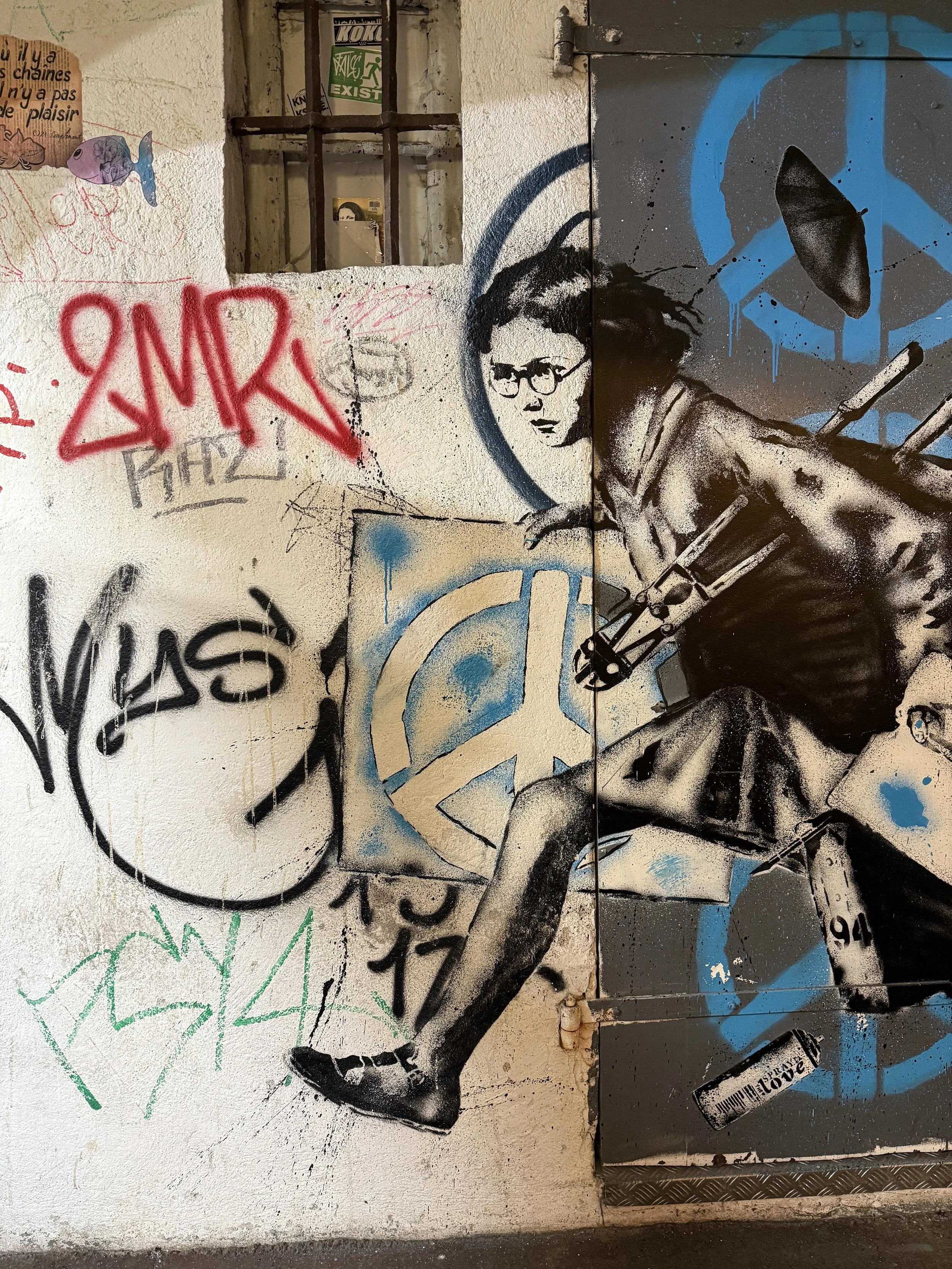 Graffiti-covered wall featuring a stencil-style figure mid-stride, blue peace symbols, and layered street tags.