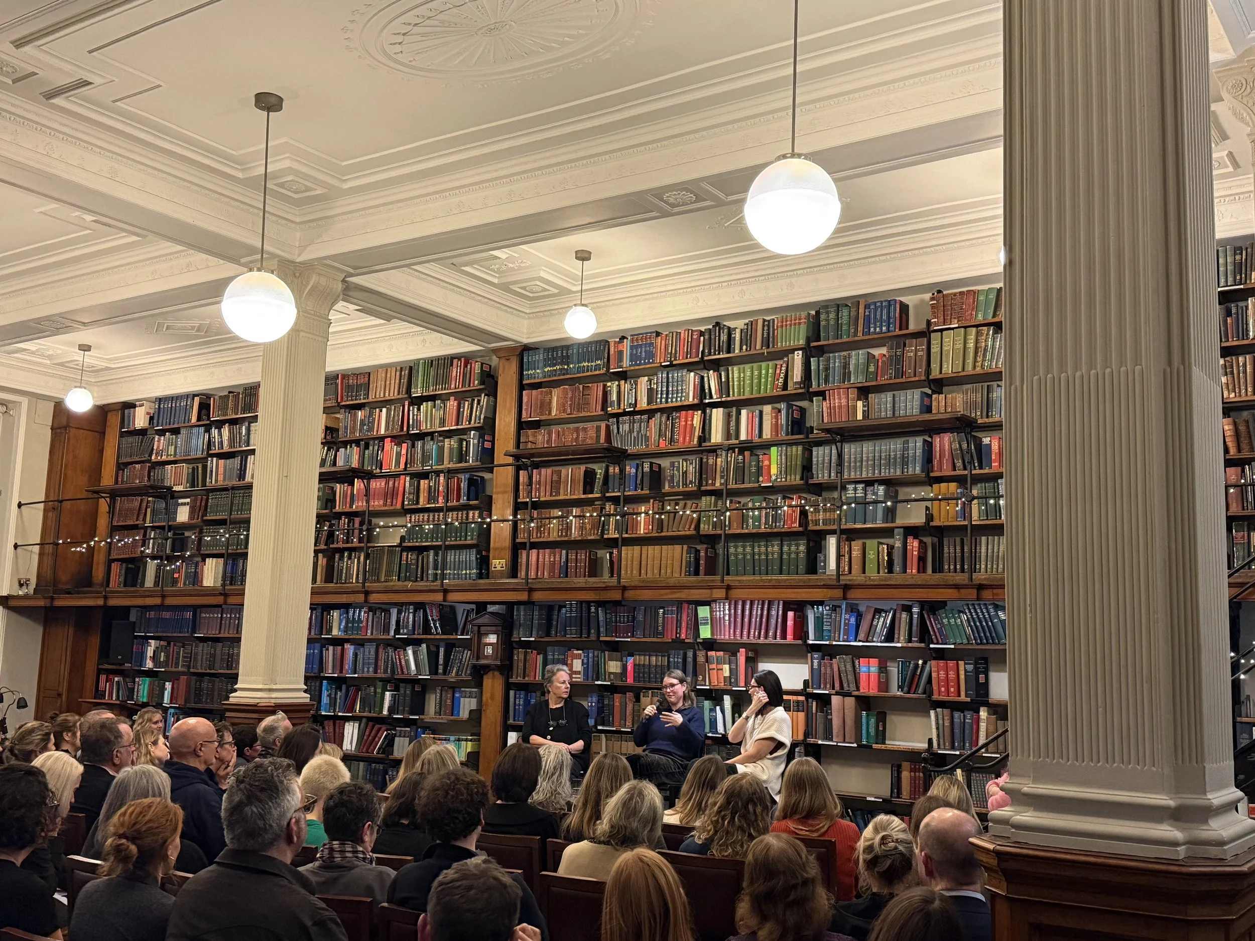 A packed evening event at the London Library, three speakers seated before a floor-to-ceiling wall of books as an audience listens.