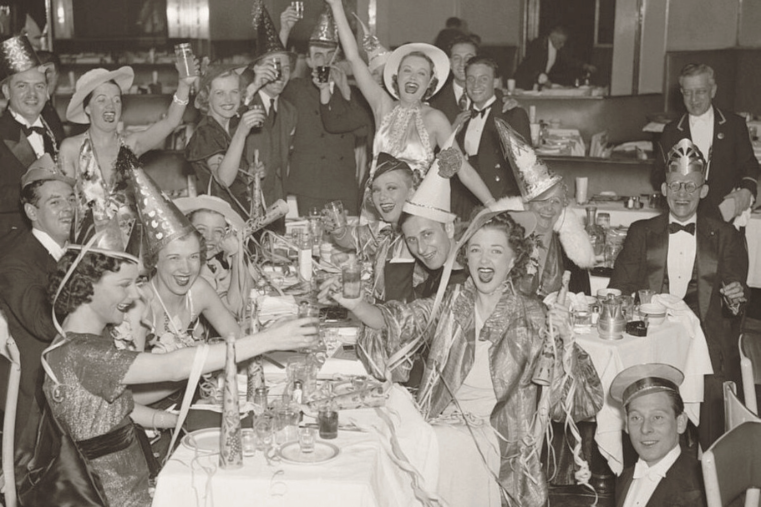 Black-and-white vintage photograph of people in party hats celebrating around a crowded table with drinks, decorations, and streamers.