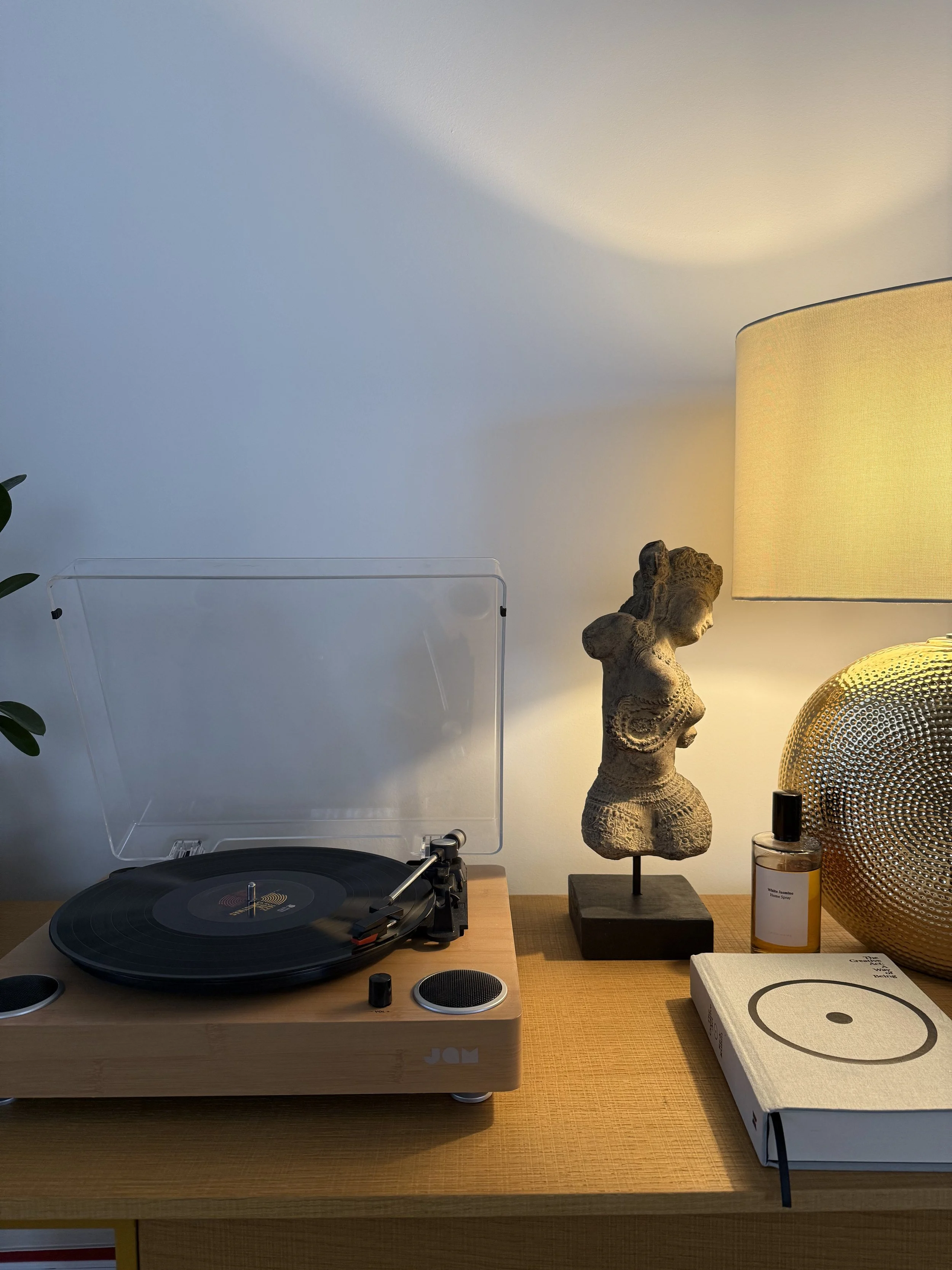 Wooden turntable with a vinyl record on a sideboard, beside a sculpted torso, glowing table lamp, perfume bottle, and book in warm, soft light.