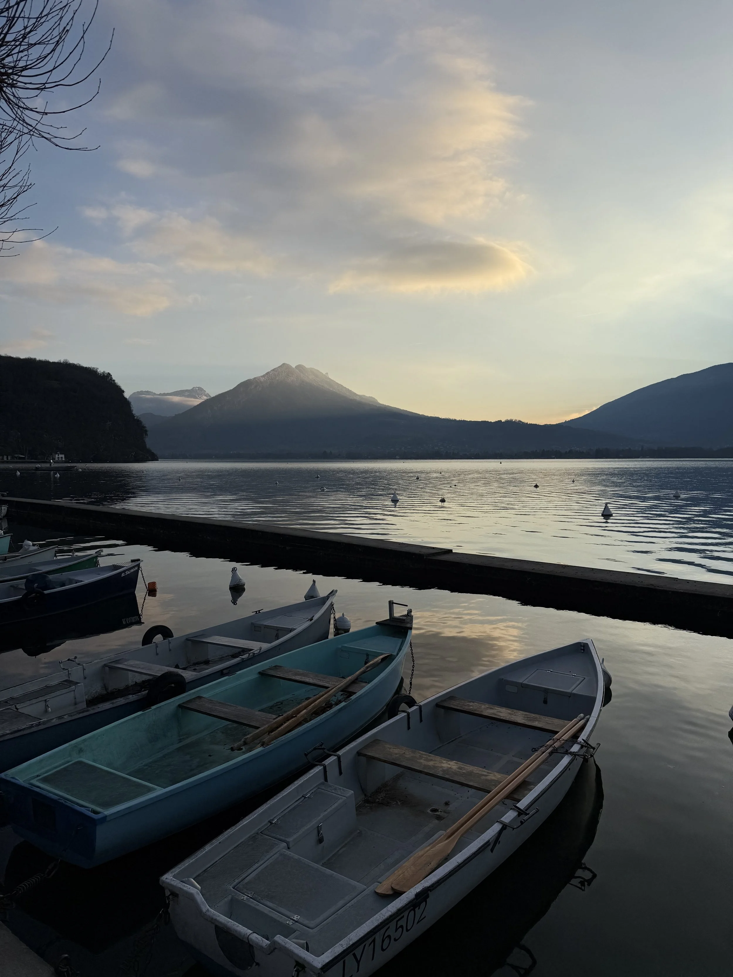 Small rowboats moored at a calm lakeside at dusk, with still water reflecting soft clouds and a mountain silhouetted in the distance.