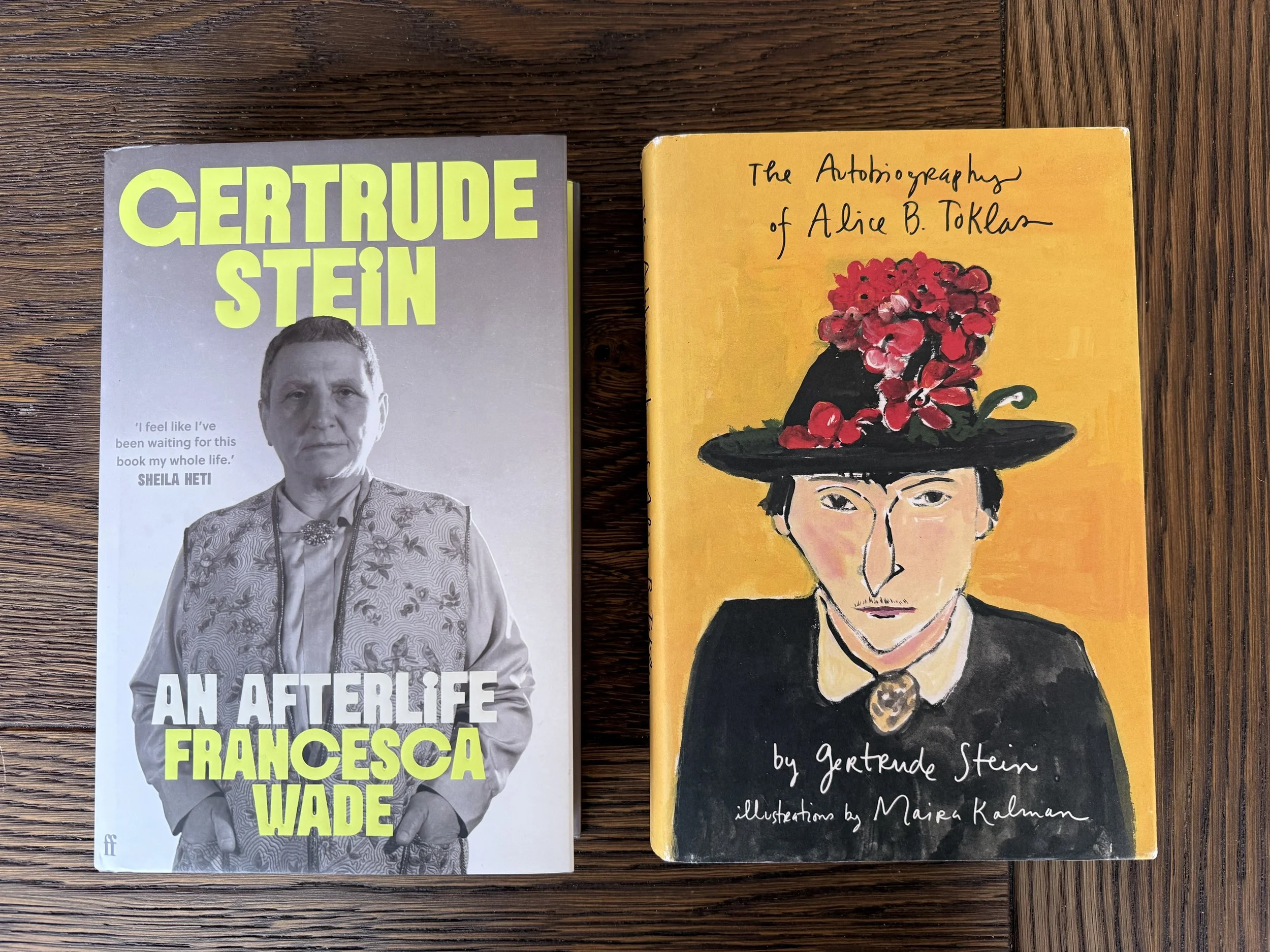 Two books on a wooden table: "Gertrude Stein: An Afterlife" by Francesca Wade, and "The Autobiography of Alice B. Toklas" illustrated by Maira Kalman.