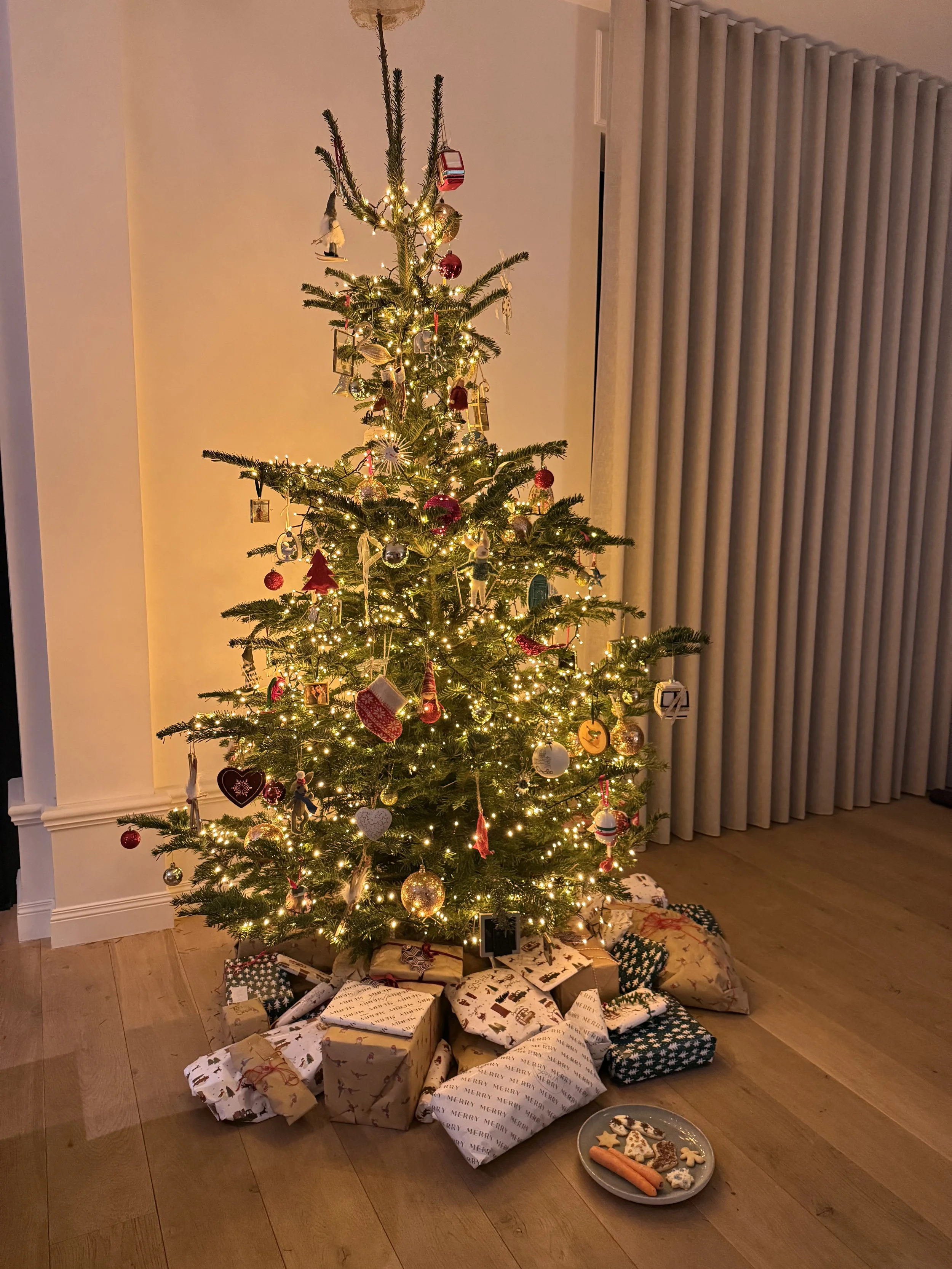 Decorated Christmas tree glowing with warm lights and ornaments, surrounded by wrapped presents on a wooden floor, with a small plate of cookies and carrots nearby.