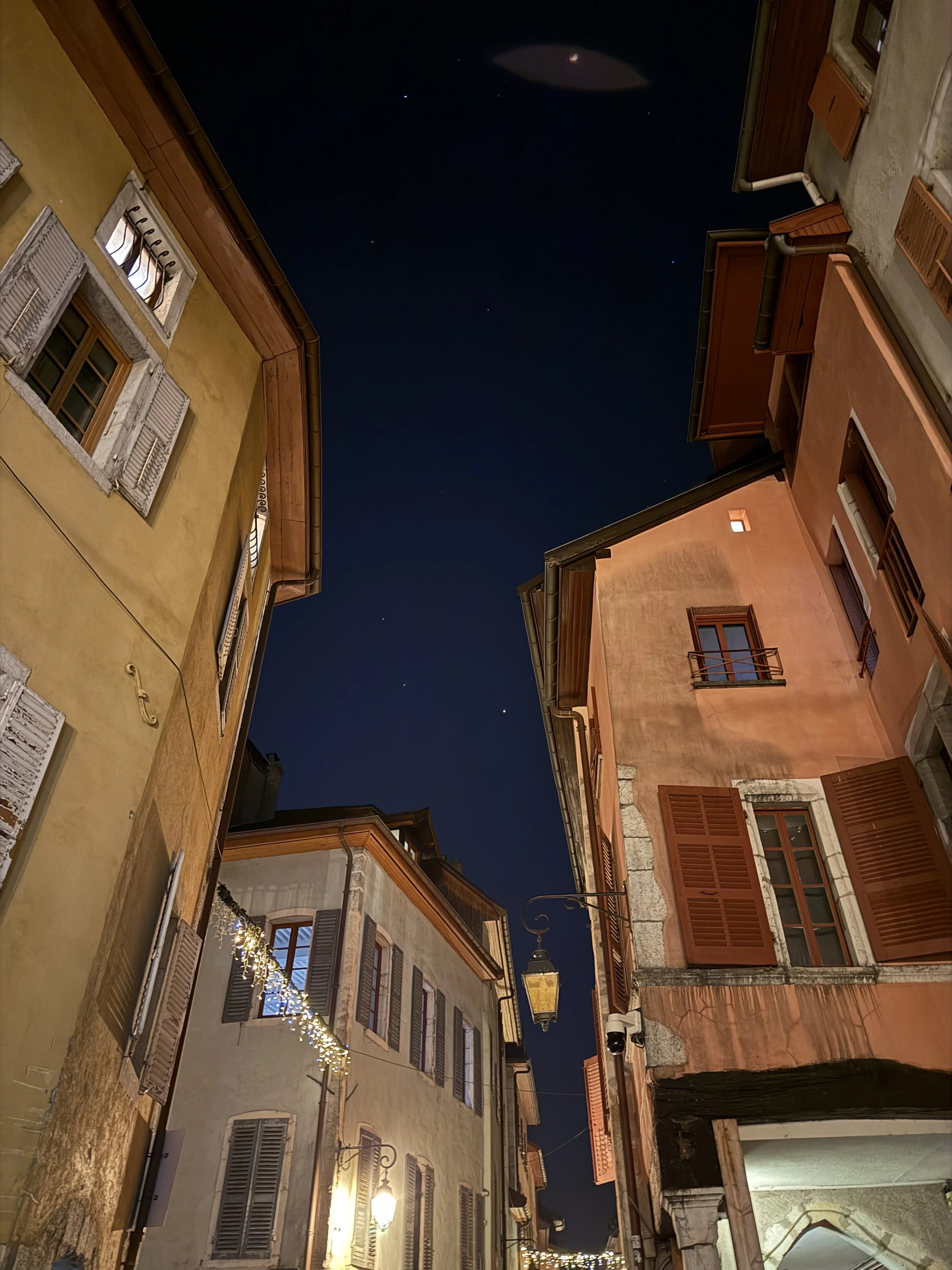 Upward view of a narrow old-town street at night, with warm-lit pastel buildings, shuttered windows, and a dark starry sky overhead.