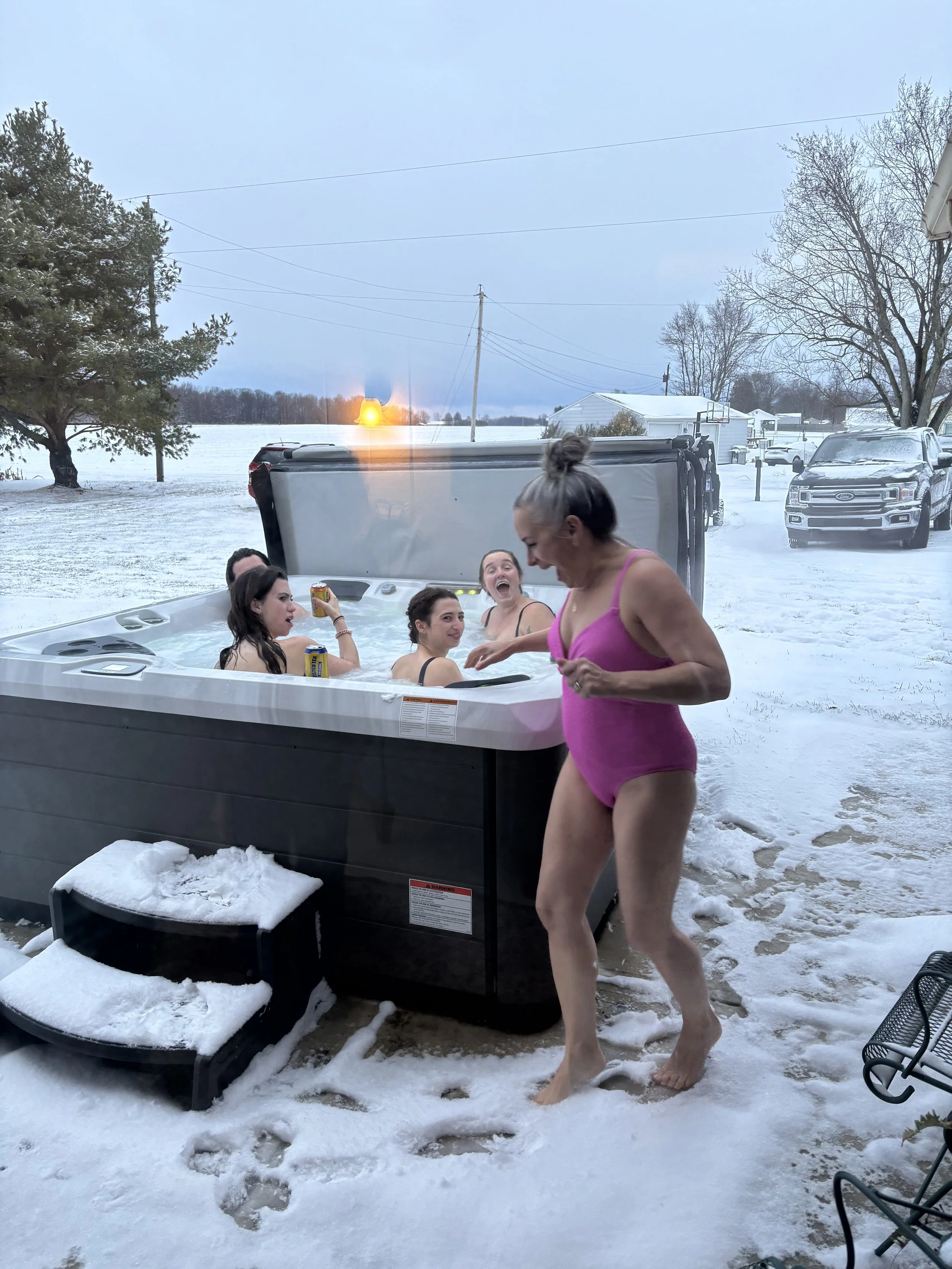 Several people sitting in an outdoor hot tub surrounded by snow, with one person stepping out onto the snowy ground.