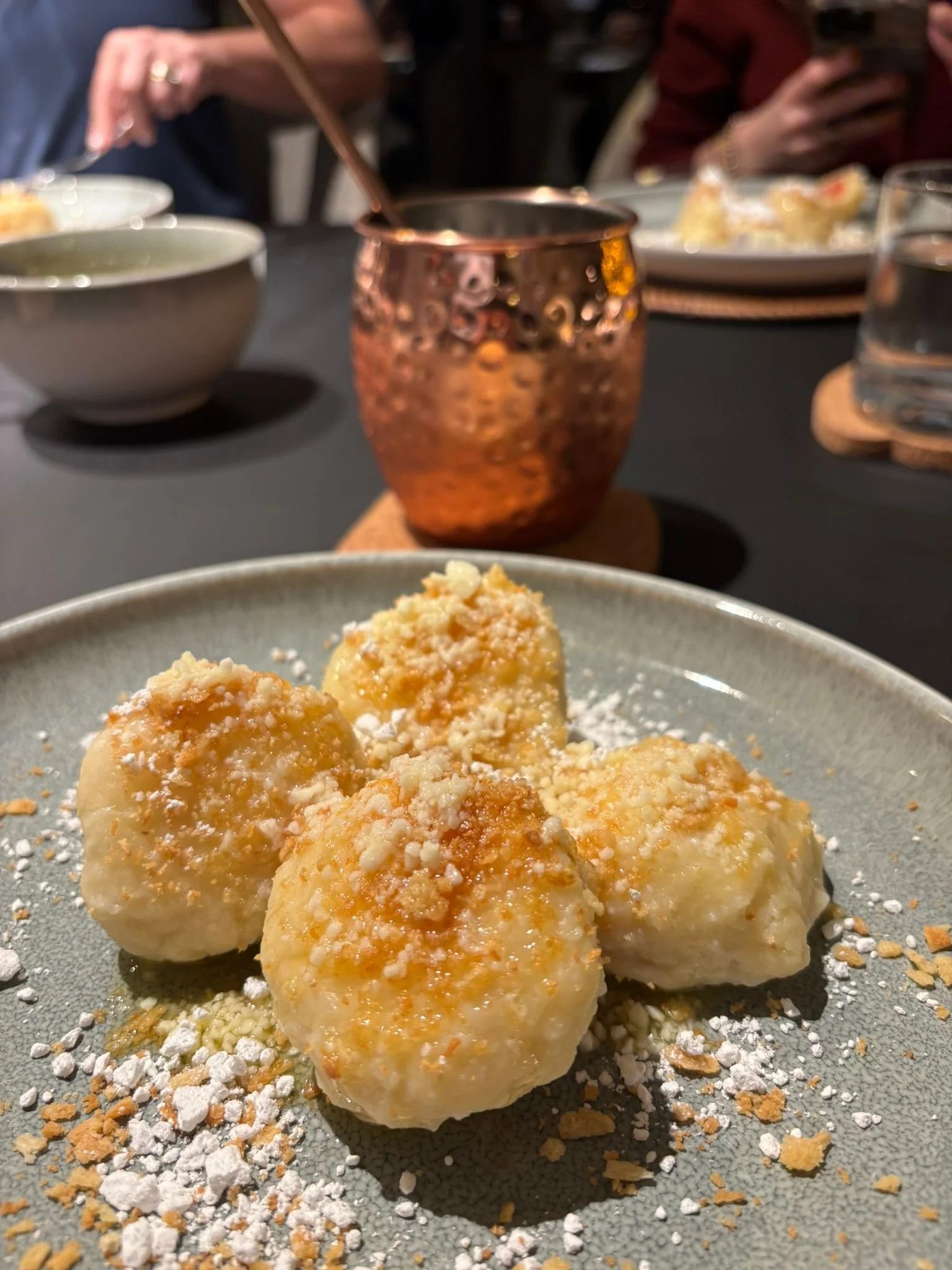 Plate of fruit dumplings dusted with sugar and crumbs on a beautiful table, with other diners and dishes softly blurred in the background.
