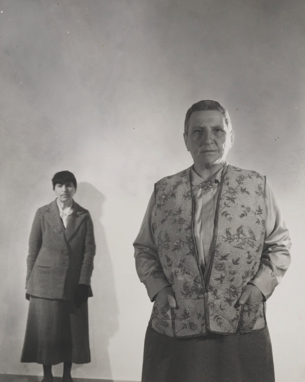 Black and white photograph of Gertrude Stein in the foreground, hands behind her back, with Alice B. Toklas standing smaller in the background — the composition saying everything the post is about.