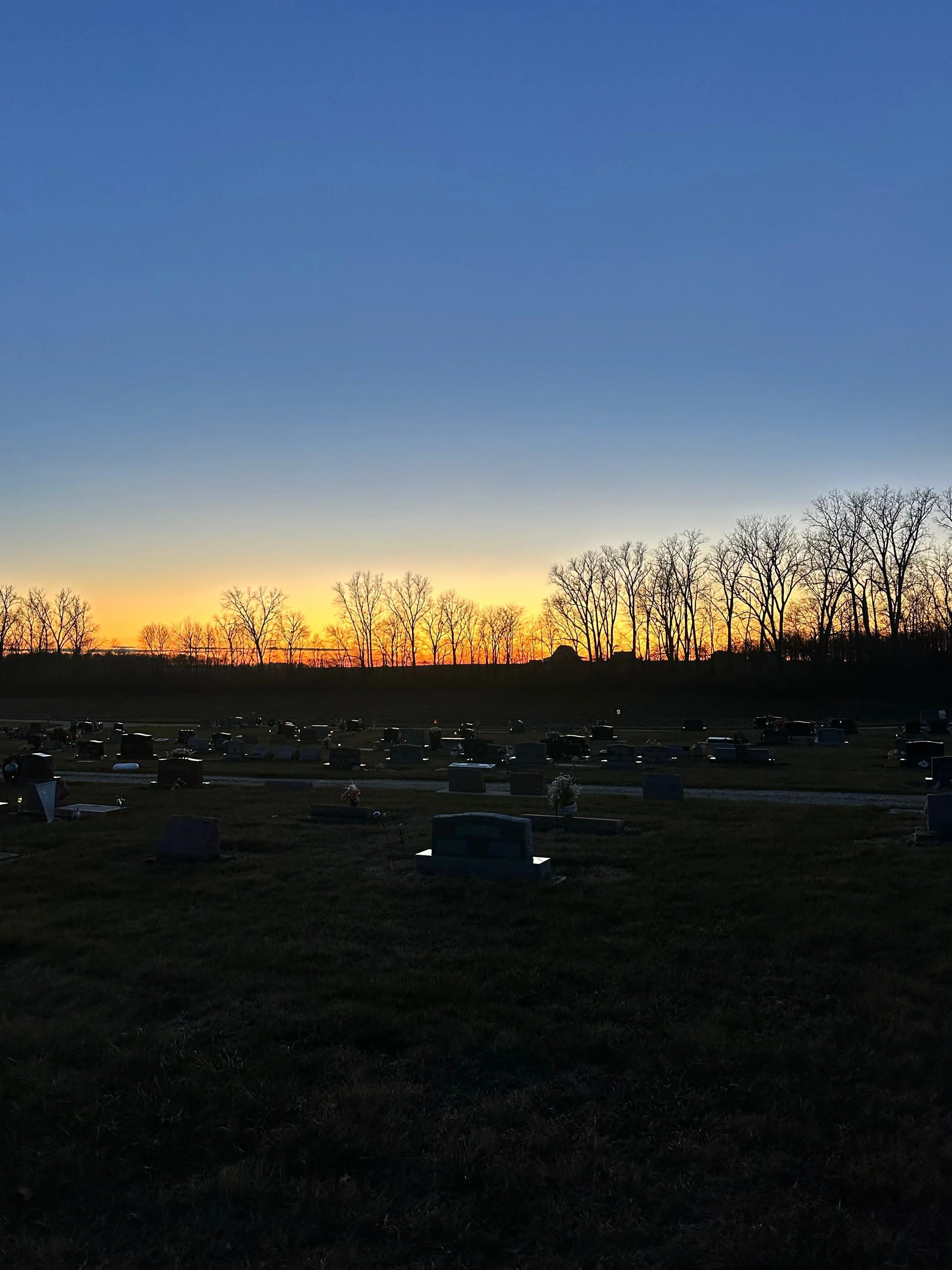 Cemetery at sunset with headstones in the foreground and bare trees along the horizon.
