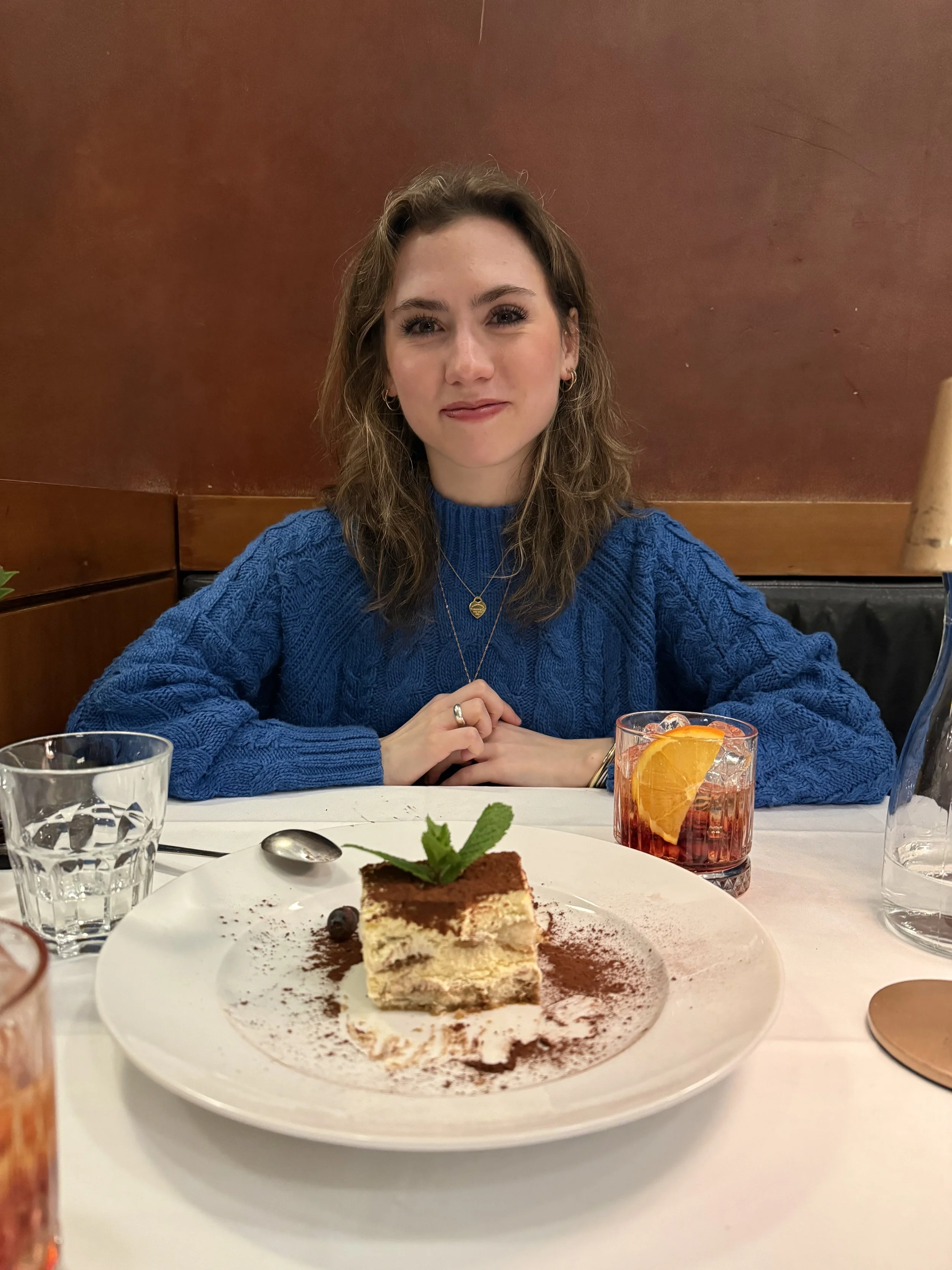 Young woman in a blue sweater sitting at a restaurant table with tiramisu and a cocktail.