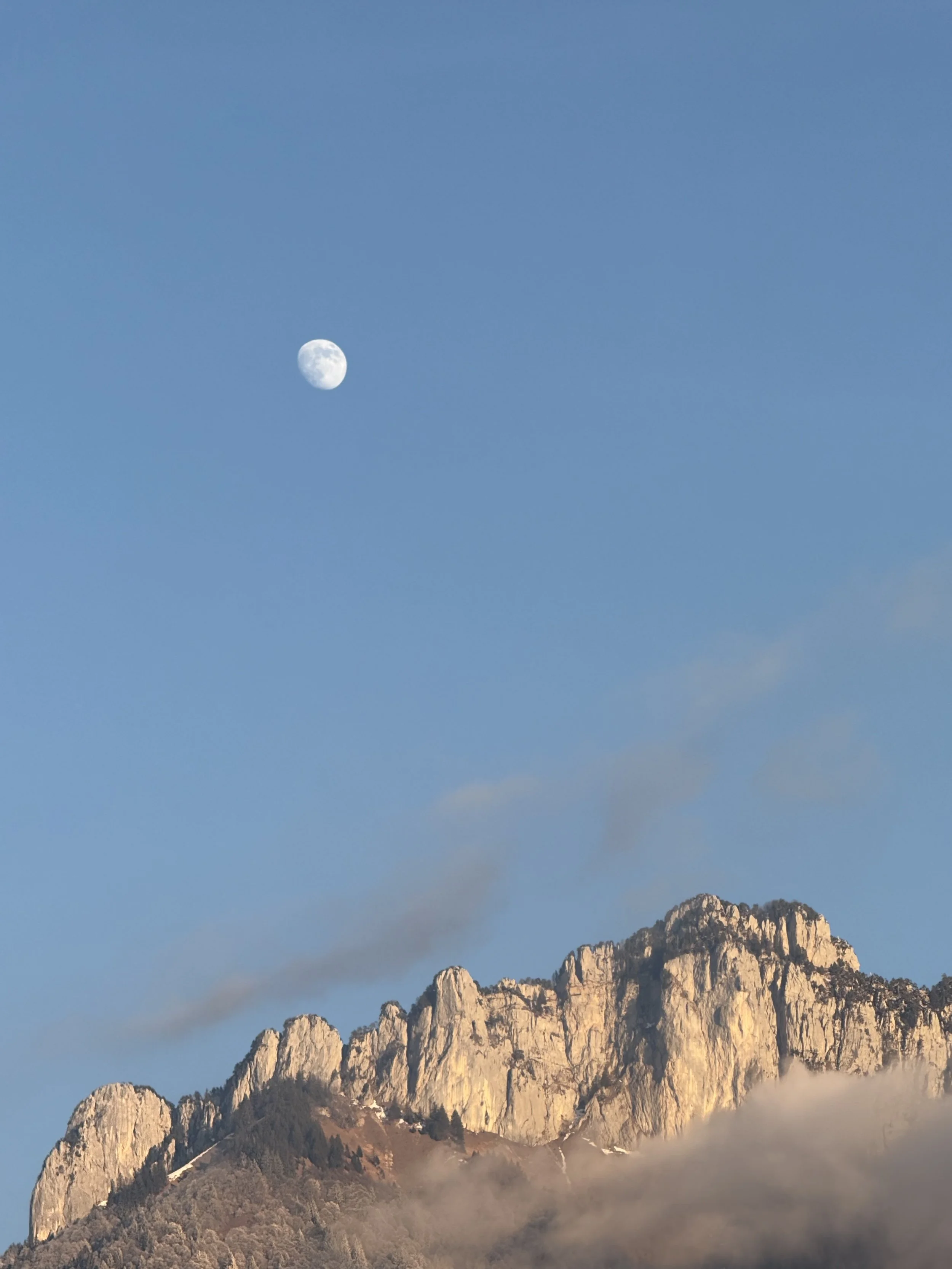Moon above a jagged limestone mountain ridge under a clear blue sky.