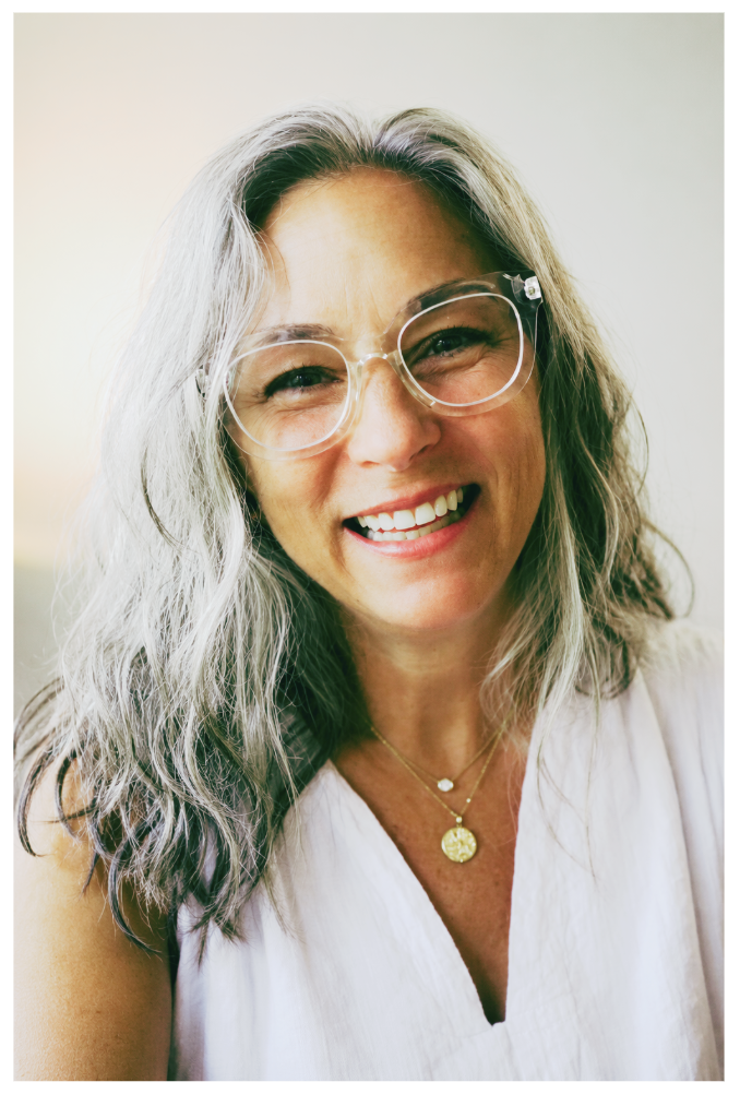 Close-up image of a smiling woman with long, wavy gray hair, wearing clear glasses and a white top, with two gold necklaces, one with a small round pendant and the other with a larger circular pendant.
