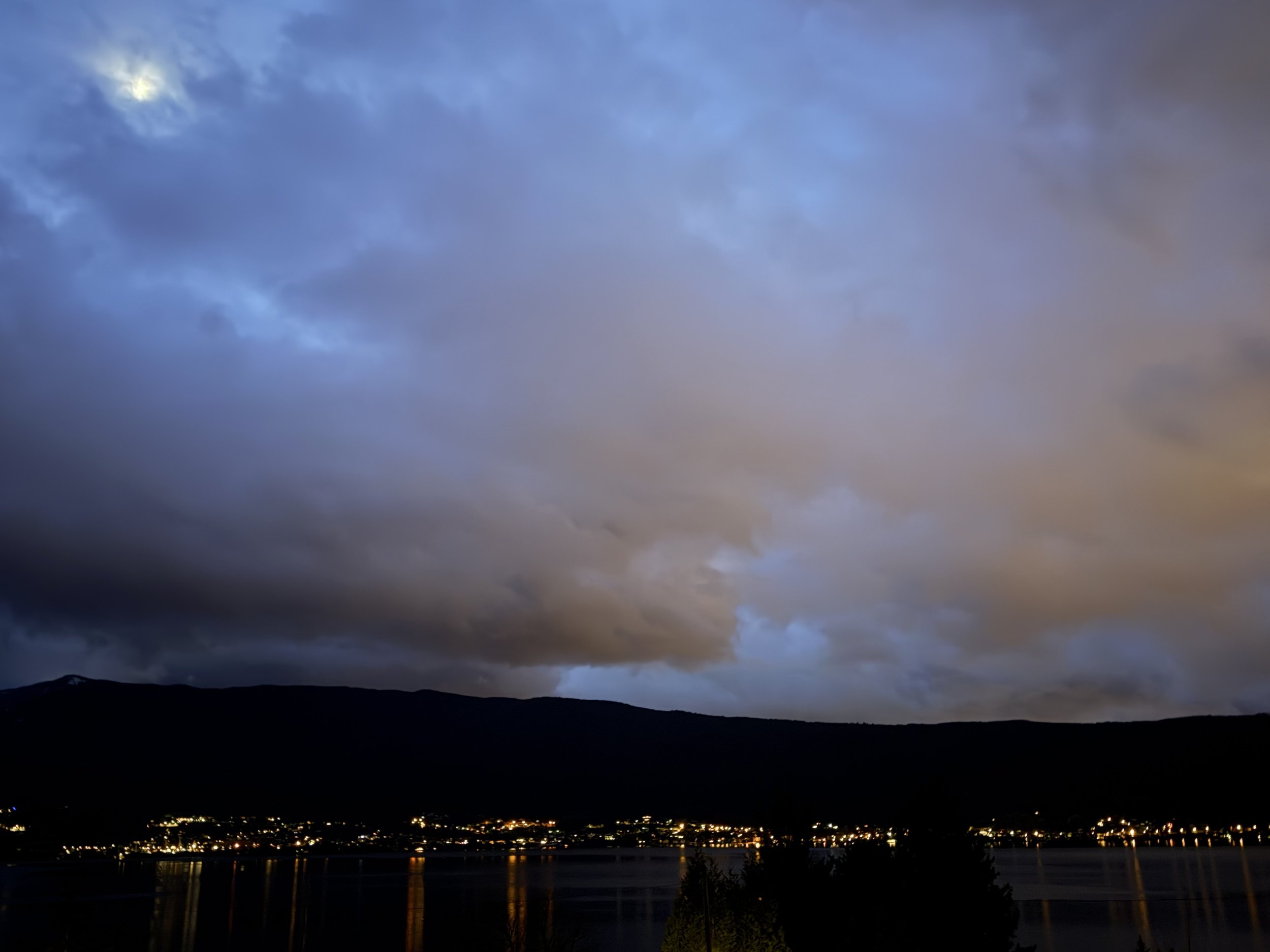 Moody dawn sky with blue and peach clouds above a dark mountain ridge, overlooking a lakeside town glowing with scattered lights reflected on the water.