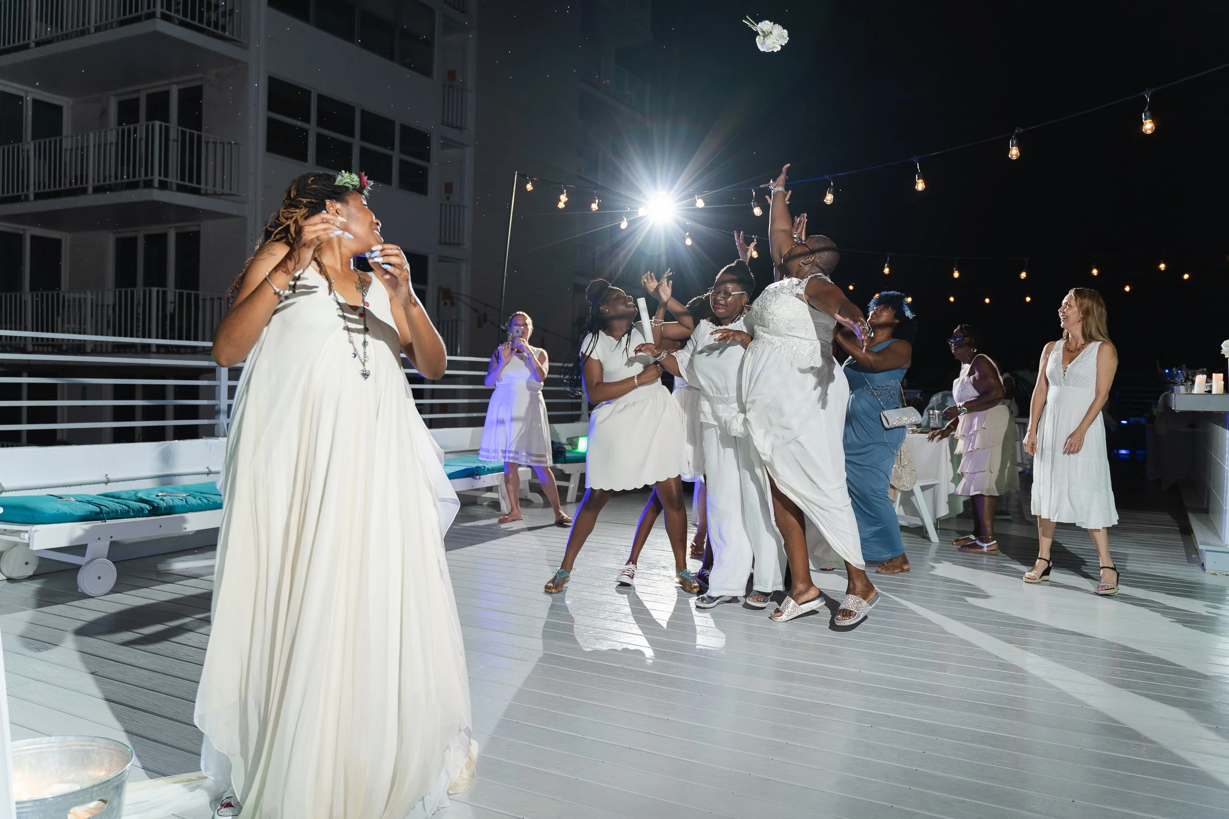 Group of women participating in a bouquet toss at an outdoor wedding reception, with a bride in the foreground holding a drink. Other guests, dressed in white and pastel dresses, are reaching for the bouquet mid-air under string lights, on a deck at 