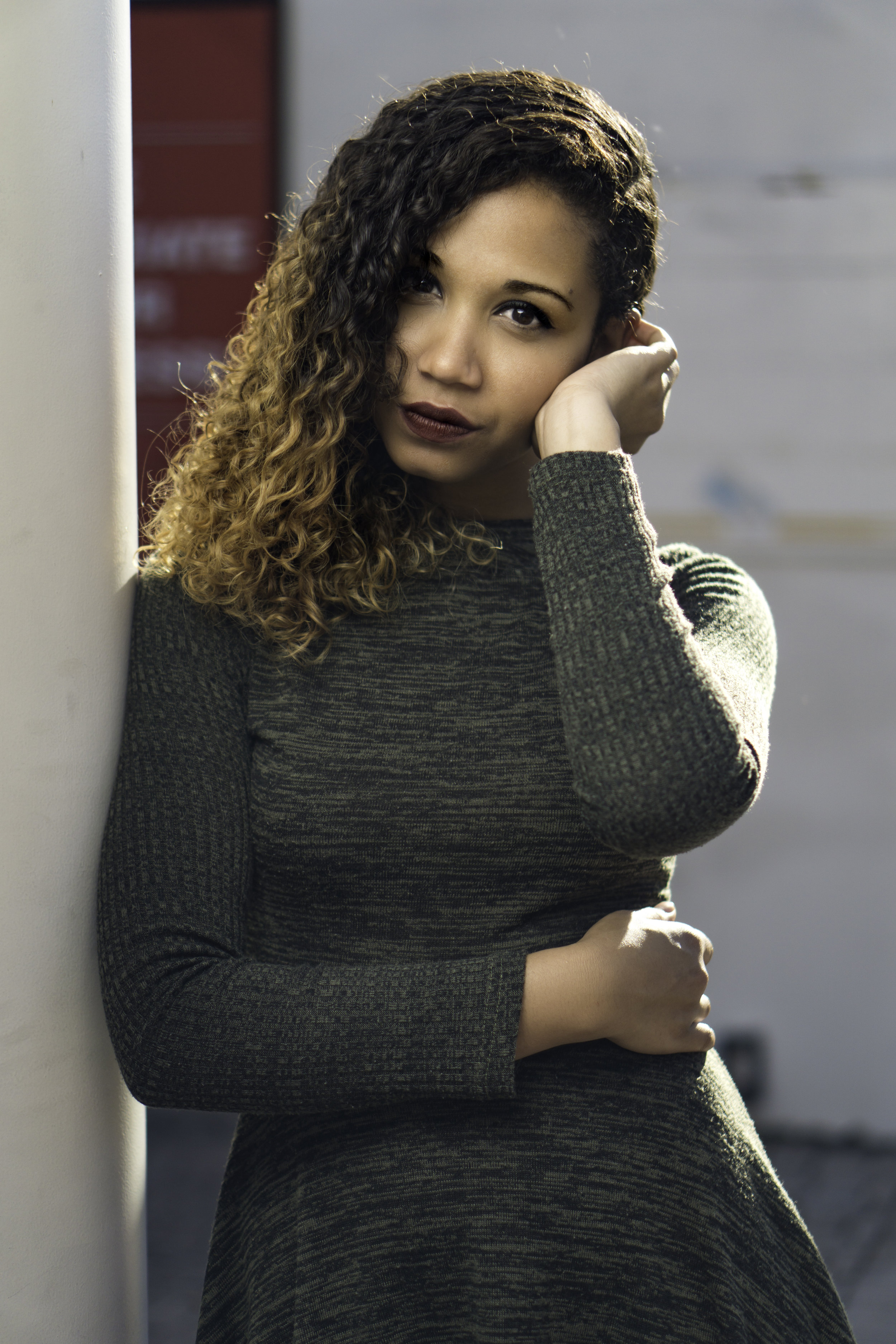 Woman with curly hair wearing a gray long-sleeved dress, leaning against a white column, looking at the camera.