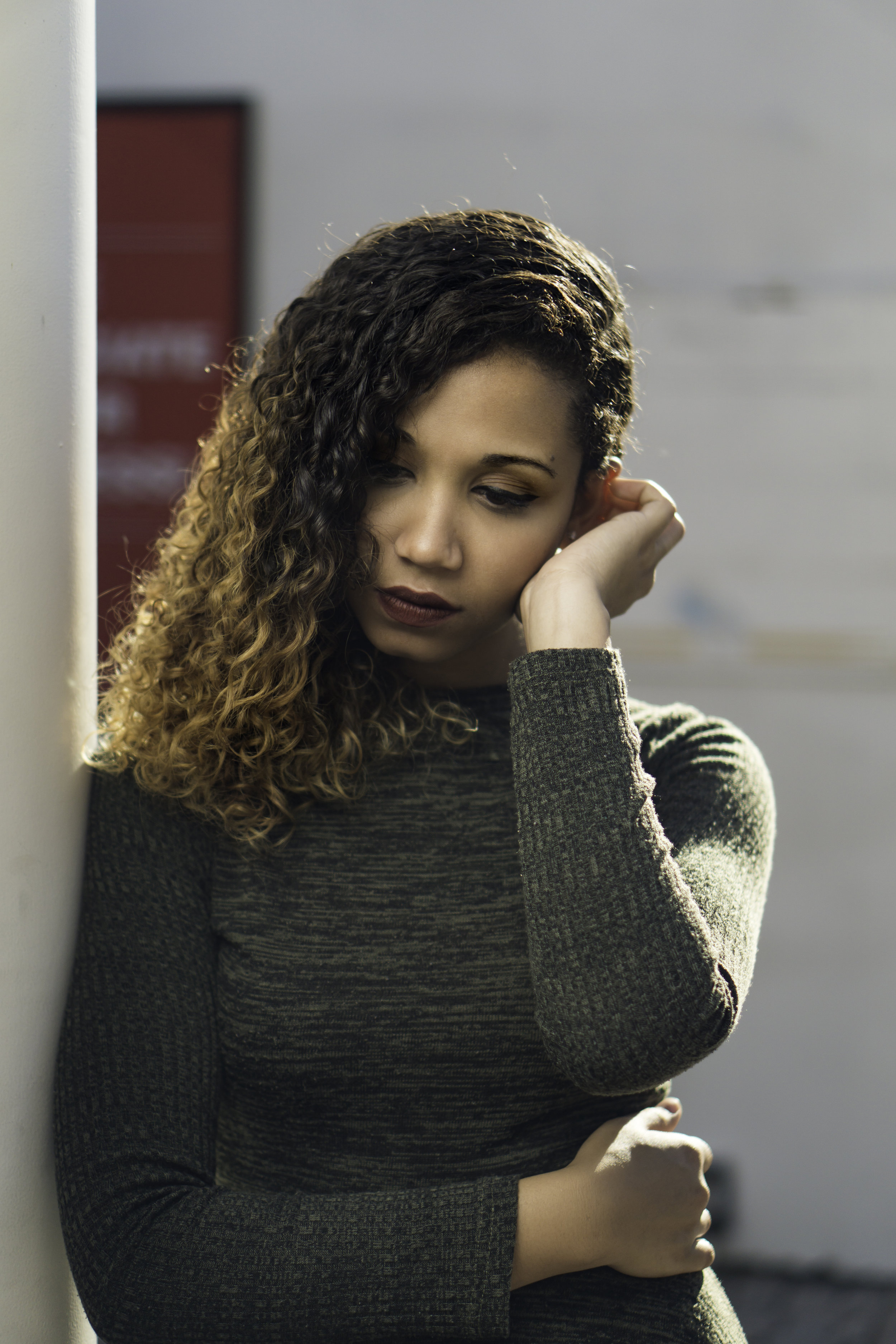 Young woman with curly hair leaning against a wall, looking thoughtful.