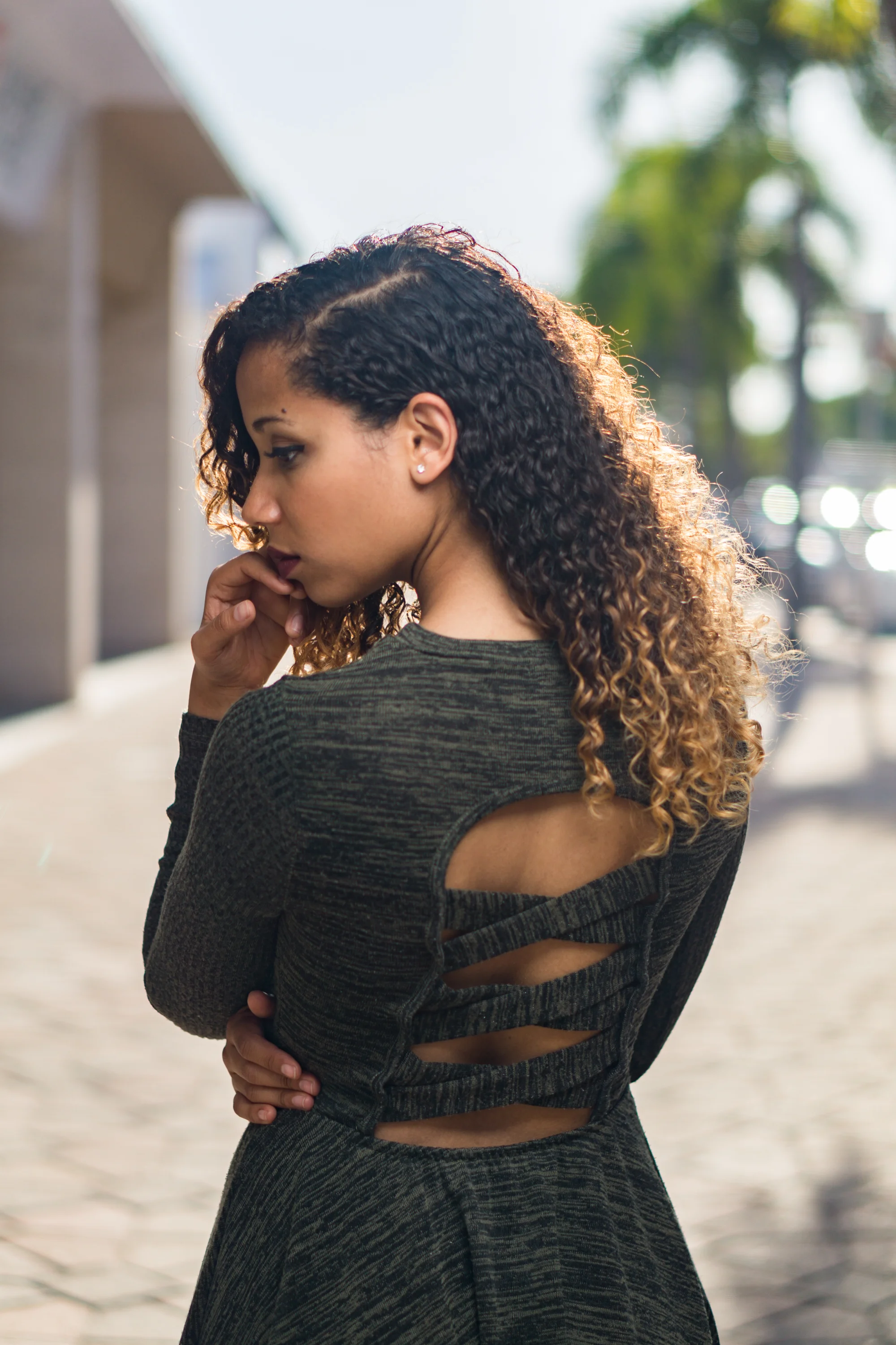Woman with curly hair in a green dress featuring cut-out back, standing outdoors with blurred palm trees in the background.